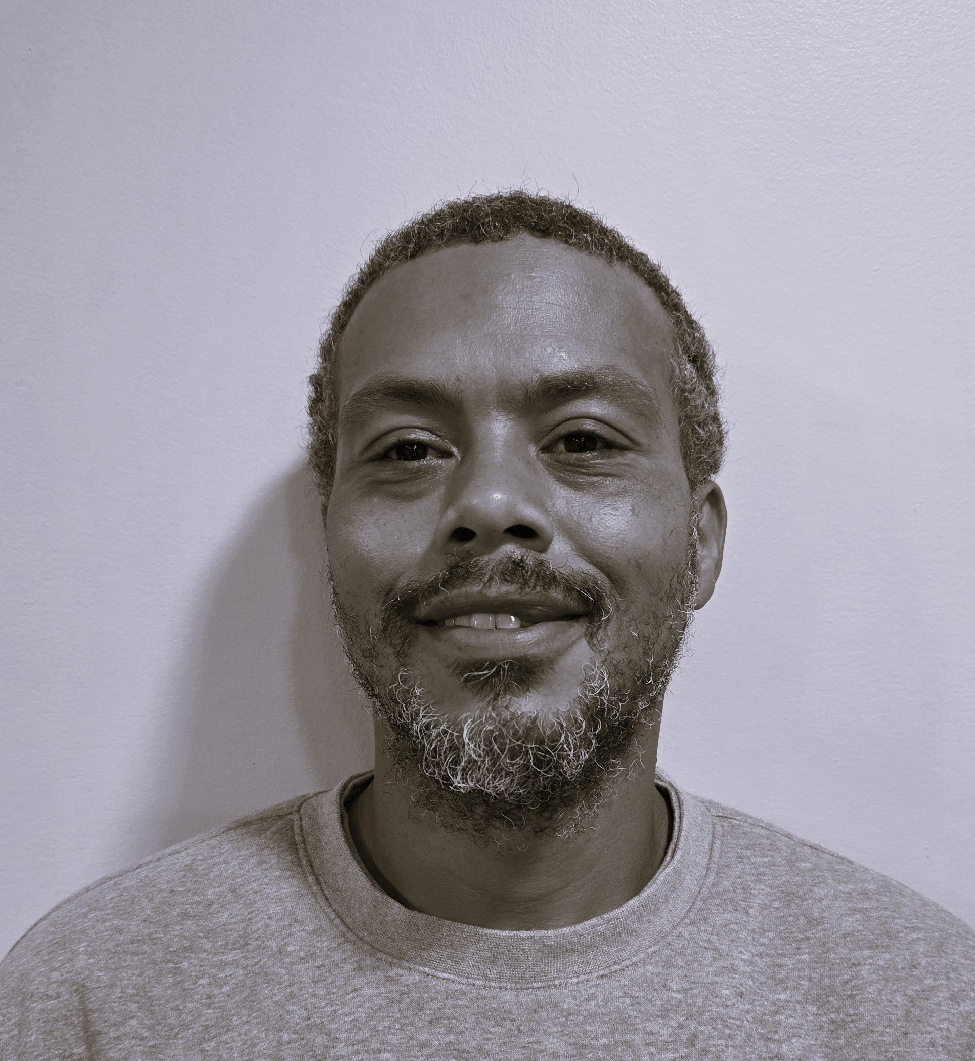 Close-up of a smiling man with gray beard and short curly hair, wearing a gray shirt, standing against a plain white wall.
