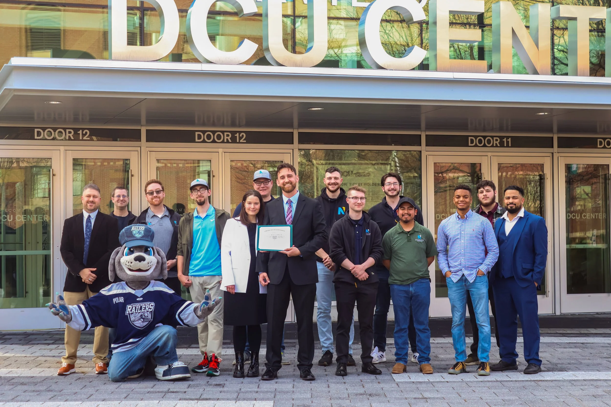 A group of people, including a mascot dressed in a hockey jersey, standing in front of a building with large glass doors. Some individuals are dressed in formal or casual attire, with one person holding what appears to be a diploma or certificate. The building has a sign with large letters that partially reads "DCU CENTER."