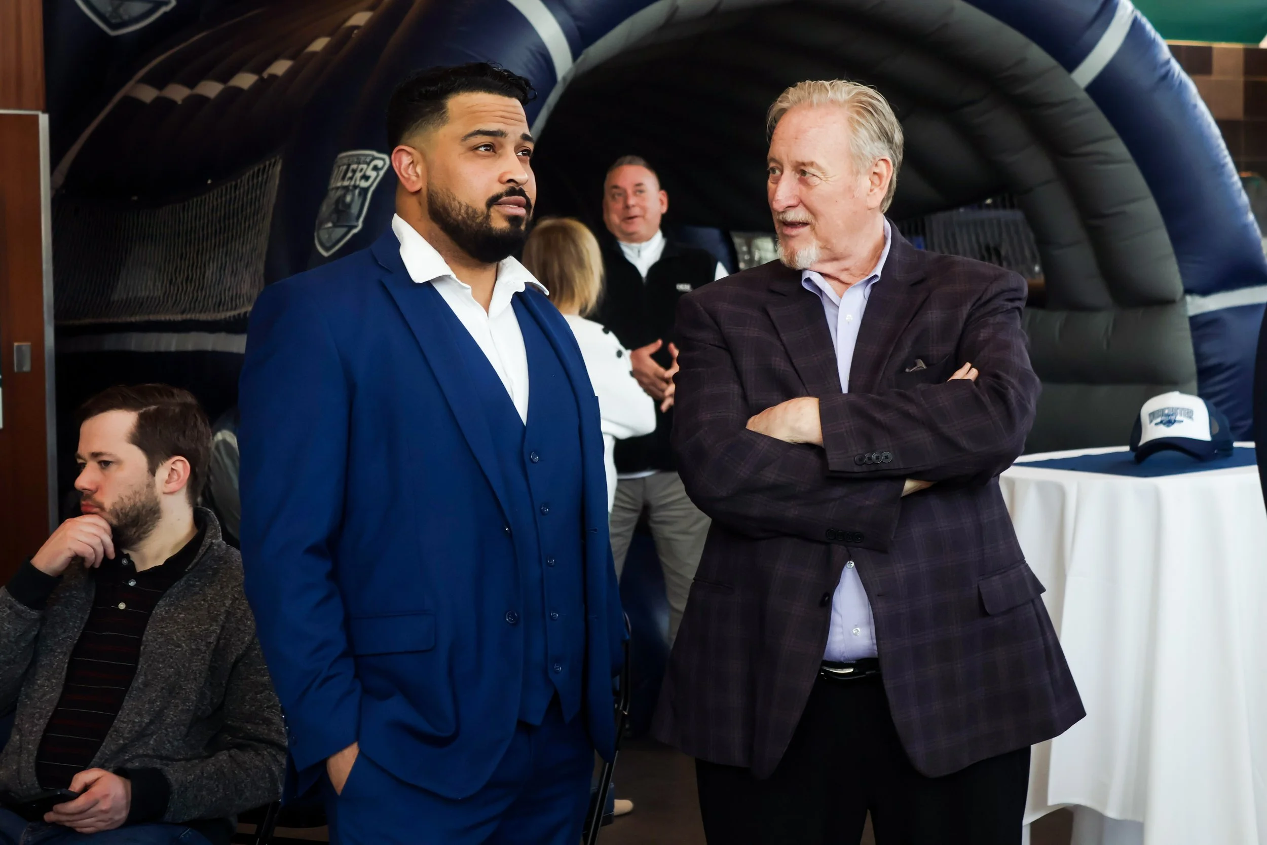 Two men engaged in conversation at a social event. The man on the left has dark hair, a beard, and is wearing a blue suit with a white shirt. The man on the right has gray hair and a beard, wearing a dark plaid blazer and white shirt. In the backgrou