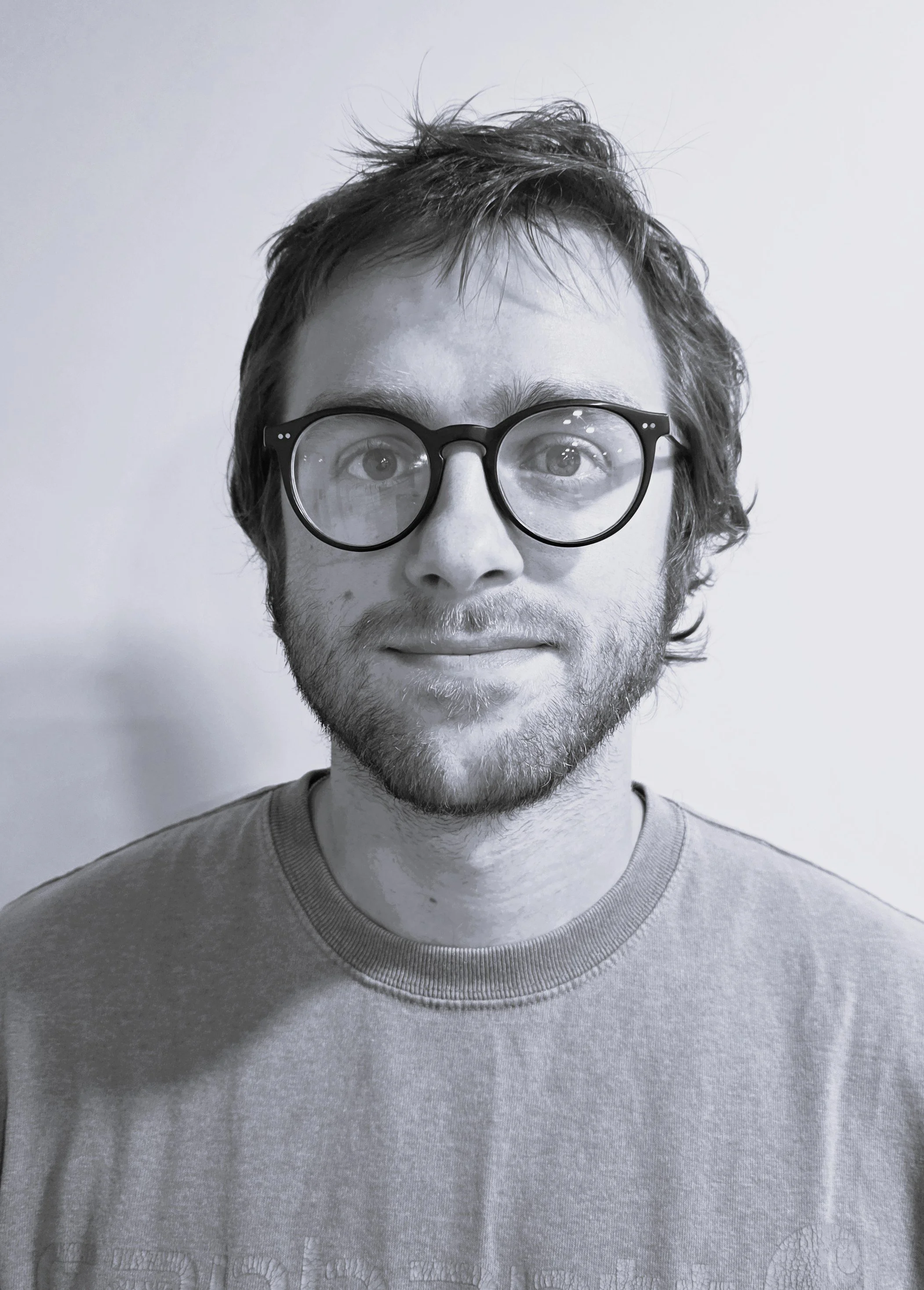 A black and white photo of a young man with wavy hair, glasses, and a slight beard, wearing a t-shirt, standing against a plain background.