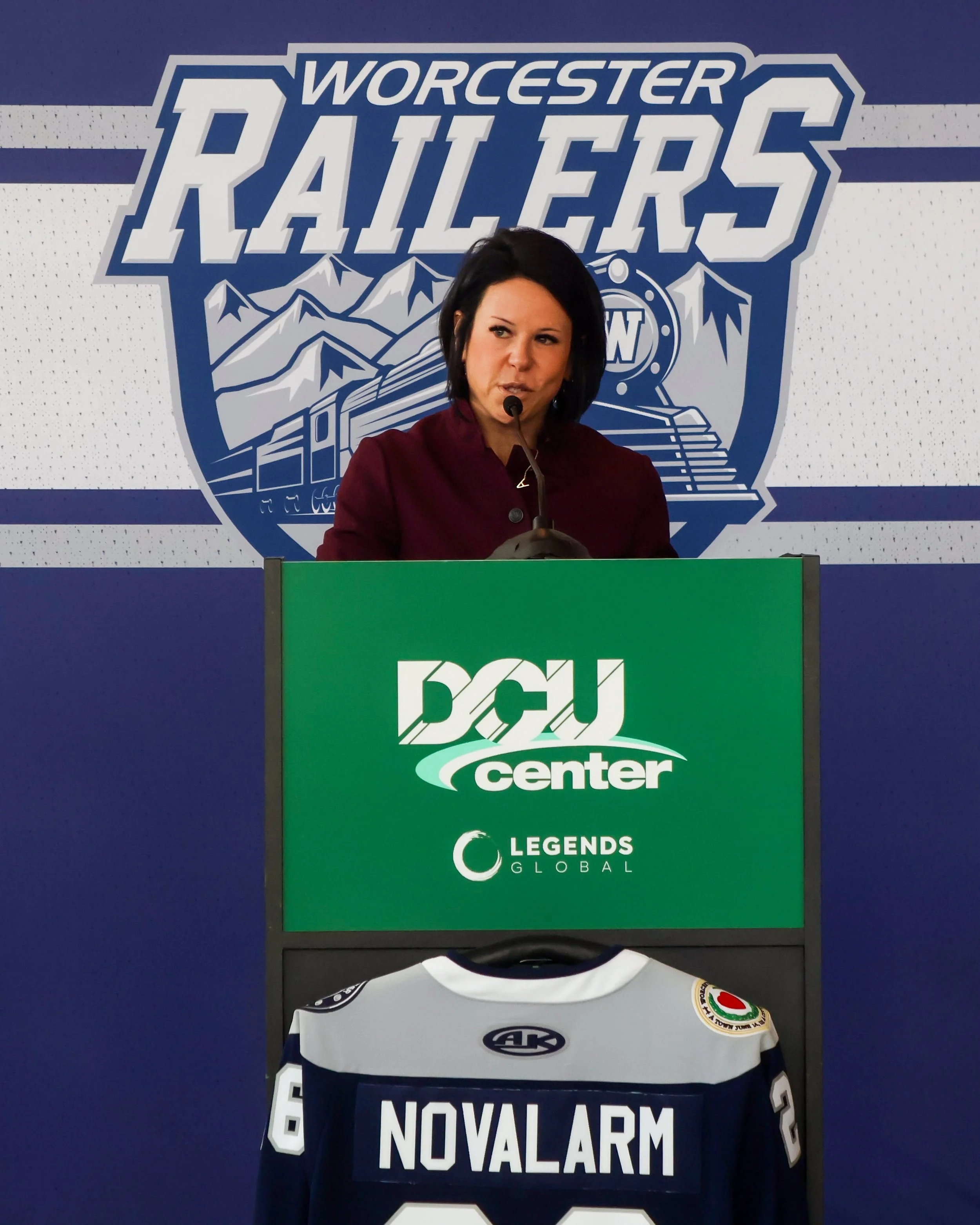 A woman standing at a podium with a microphone, with a backdrop that says 'Worcester Railers' and has a mountain and train graphic. She is wearing a maroon jacket.