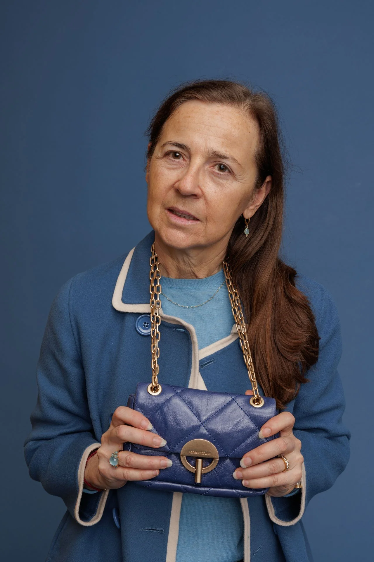 Une femme avec des cheveux bruns longs portant un manteau bleu tenue d'une femme d'âge moyen tenant un sac à main bleu avec une chaîne en or contre un fond bleu.