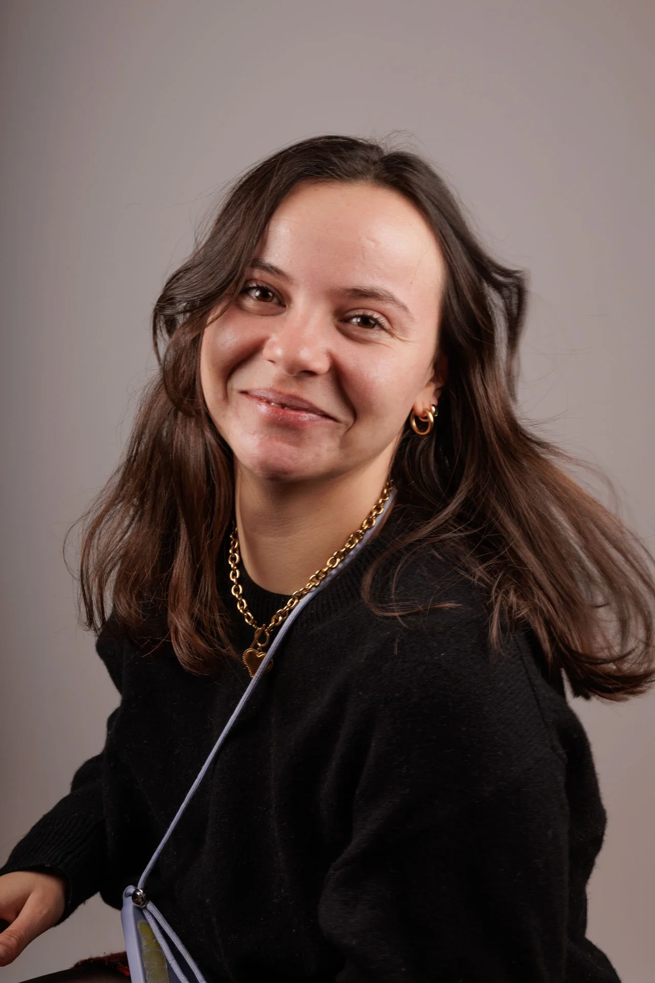 Portrait d'une femme avec des cheveux bruns, portant des bijoux en or, un pull noir, et souriant légèrement, avec un fond gris.