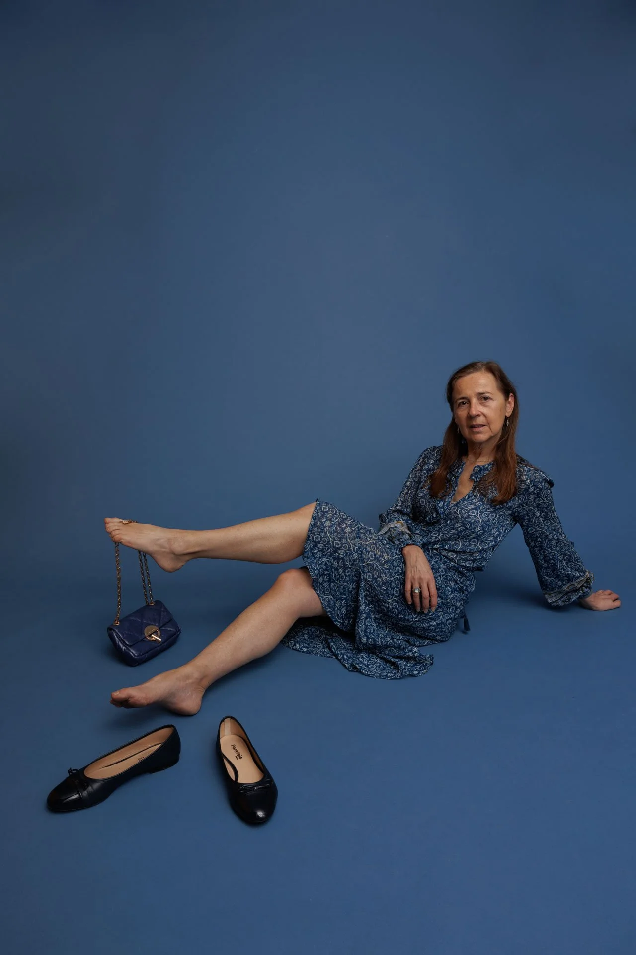 Femme âgée portant une robe bleue, assise par terre avec des chaussures noires, un sac à main et une bague, contre un fond bleu.