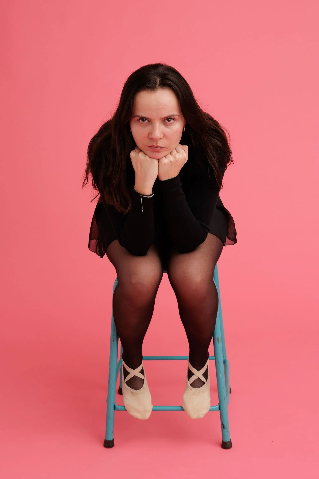 Jeune femme assise sur une chaise en traçant une pose de défi, portant un vêtement noir, des collants noirs et des chaussures beige avec des lacets croisés, contre un fond rose.