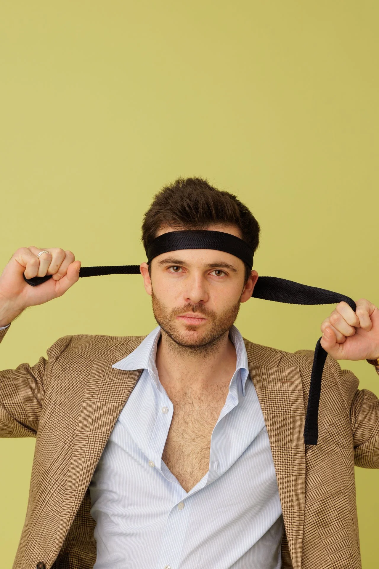 Un homme avec un blazer marron, une chemise blanche légèrement ouverte, porte un bandeau noir sur la tête et tient une corde noire derrière la tête.