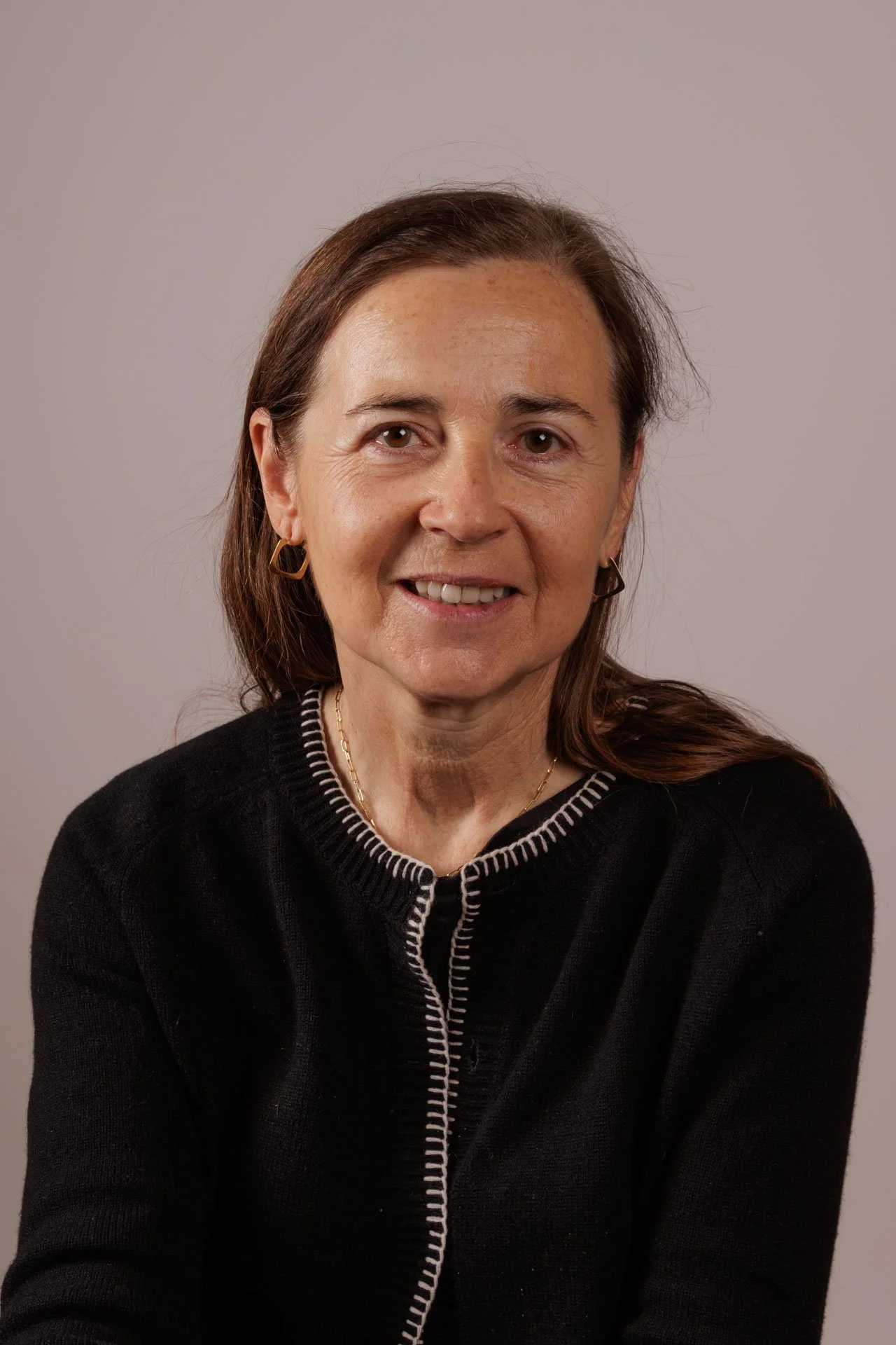 Portrait d'une femme avec des cheveux bruns, portant un pull noir avec des coutures blanches, portant des boucles d'oreilles dorées et un collier doré, contre un fond neutre.
