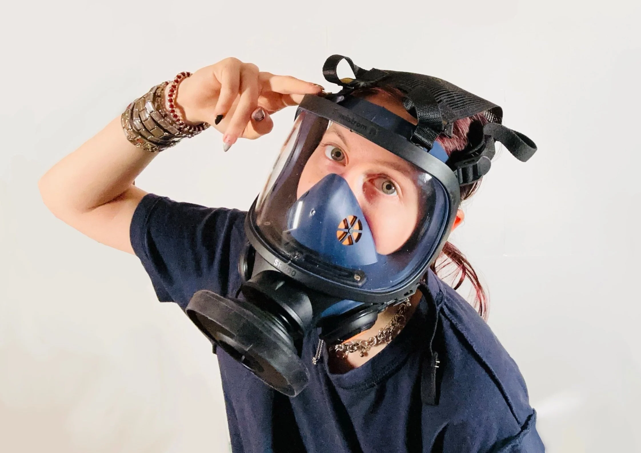 Junge Person trägt eine Schutzmaske und eine Schutzbrille, zeigt ein peace-Zeichen mit der Hand, trägt Schmuck und ein schwarzes T-Shirt, steht vor weißem Hintergrund.