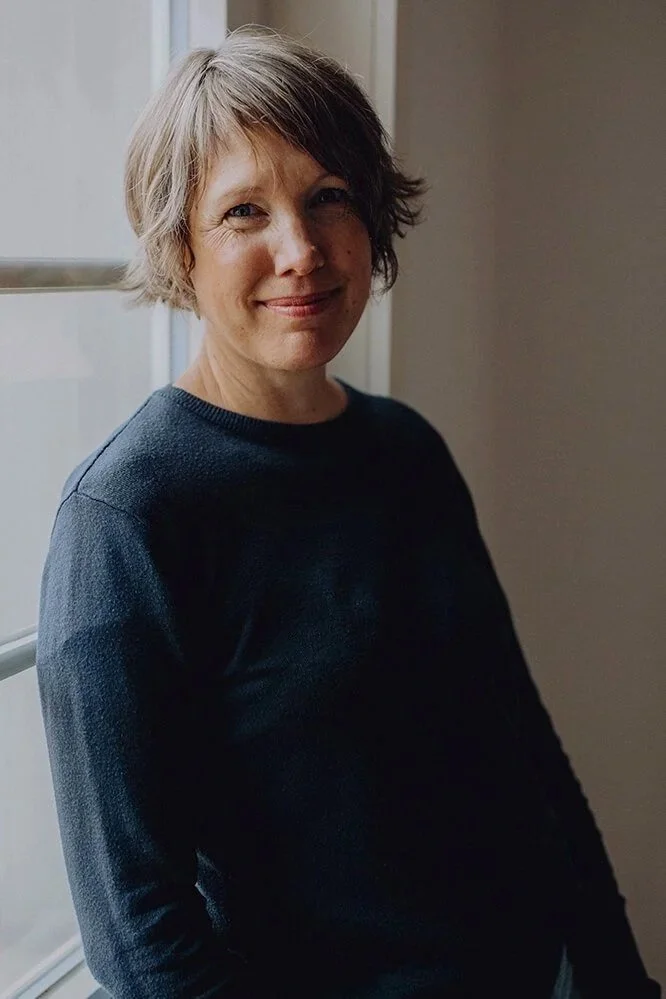 Portrait von Sara Sommer sie schaut freundlich in die Kamera hat einen blauen Pullover an und steht angelehnt am Fenster