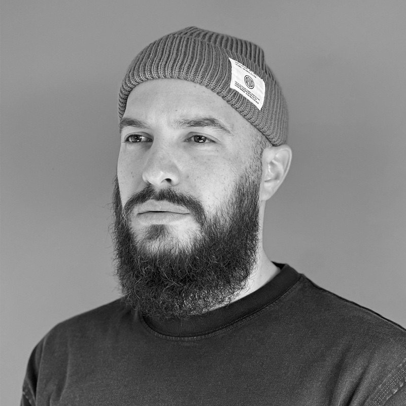 Portrait of a bearded man wearing a beanie and a sweater, looking serious against a plain background.