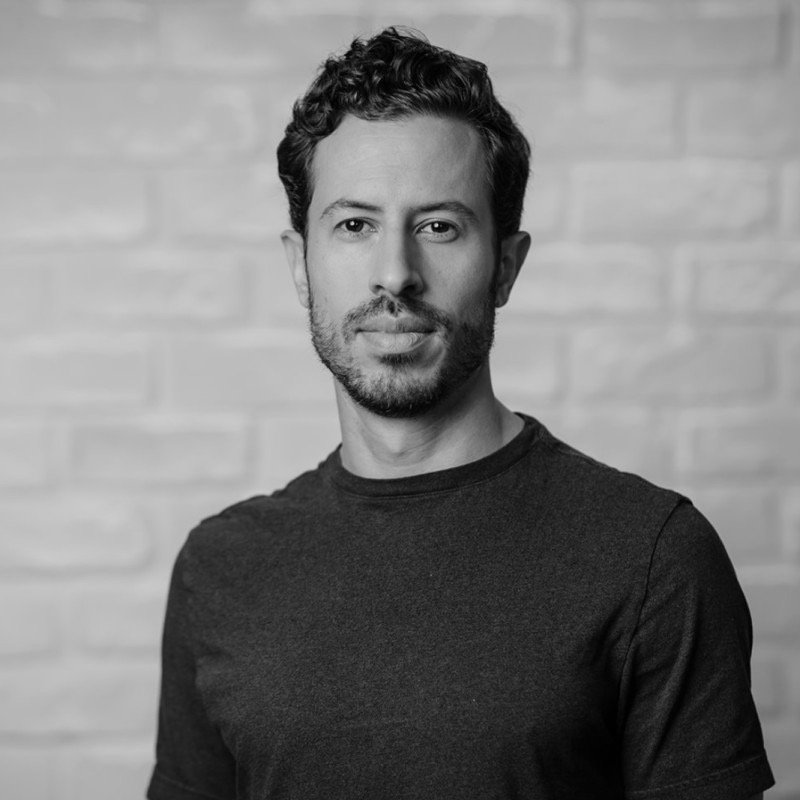 Black and white portrait of a man with a beard and curly hair, wearing a dark t-shirt, standing in front of a brick wall.