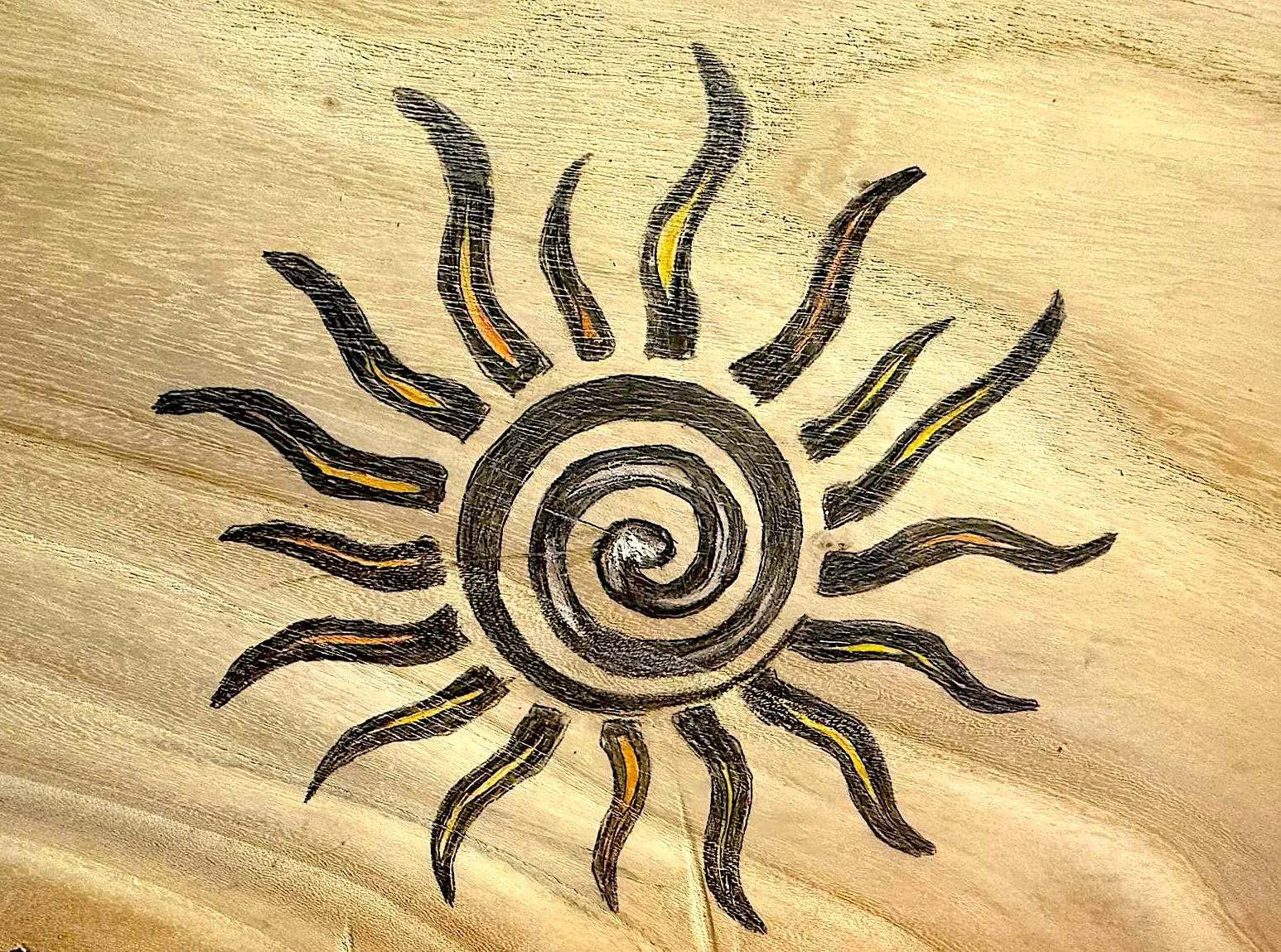 Disegno di un sole stilizzato con un vortice centrale e raggi ondulati, di colore nero e giallo su sfondo di legno chiaro.