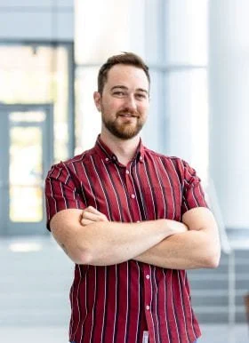 A young man with a beard and short hair, wearing a red and black striped shirt, standing indoors with arms crossed and smiling.