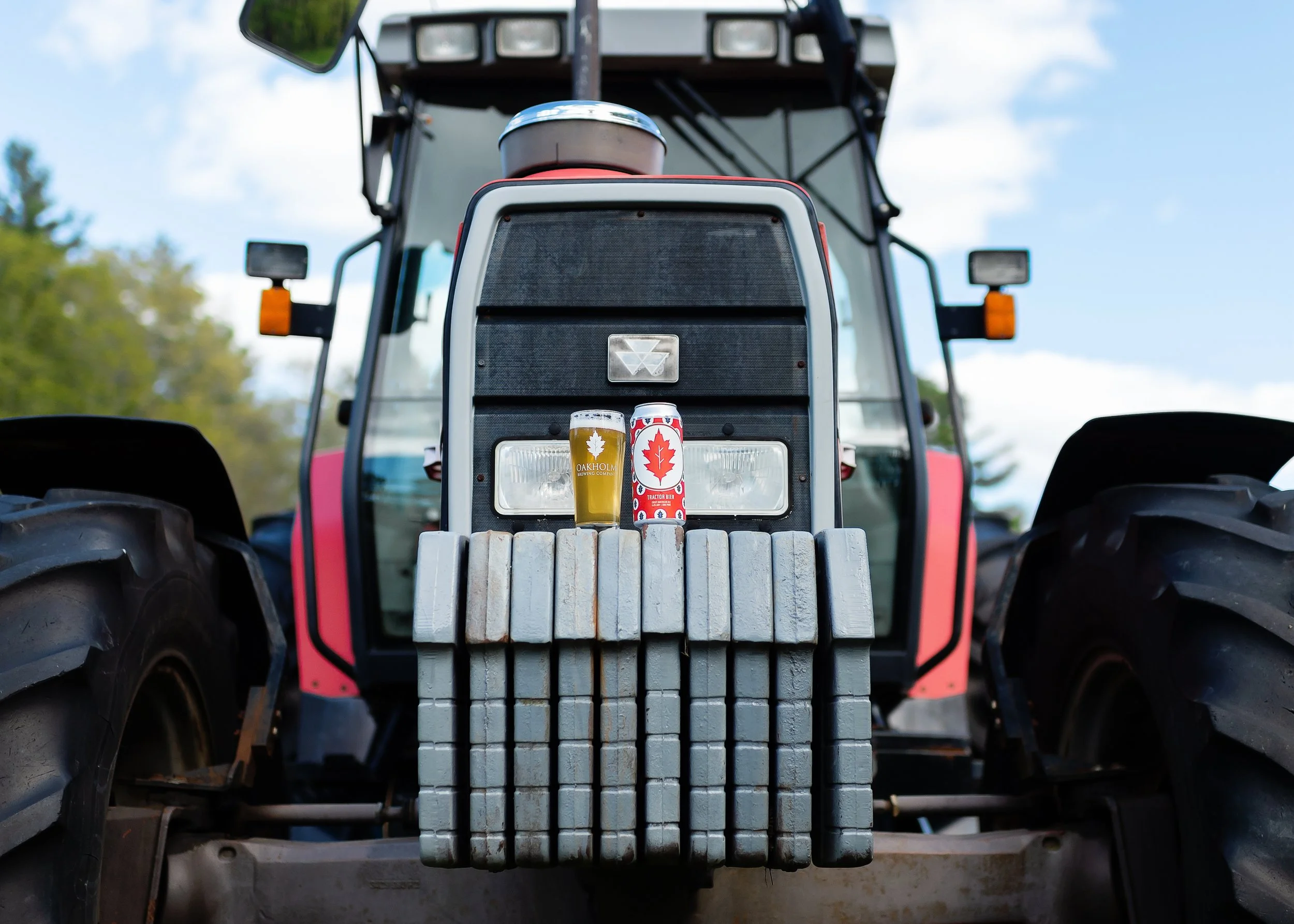 Front view of a tractor with two cans on its front grille, one containing beer and the other a beverage in a red and white maple leaf-themed can, outdoors with a blue sky and green trees.