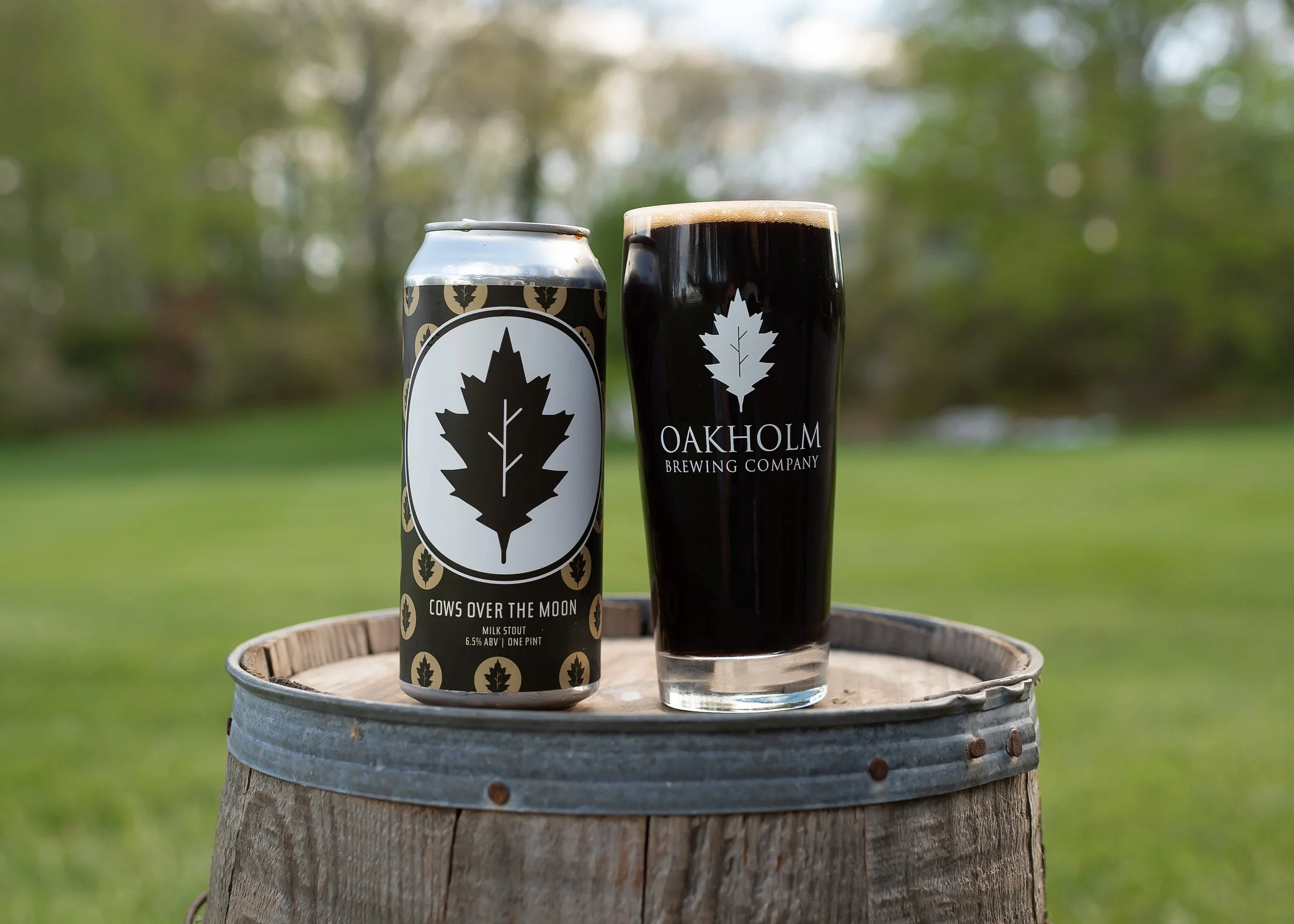A can of Cows Over the Moon milk stout and a glass of dark beer, both with similar leaf logos, on a wooden barrel outdoors with a grassy field and trees in the background.