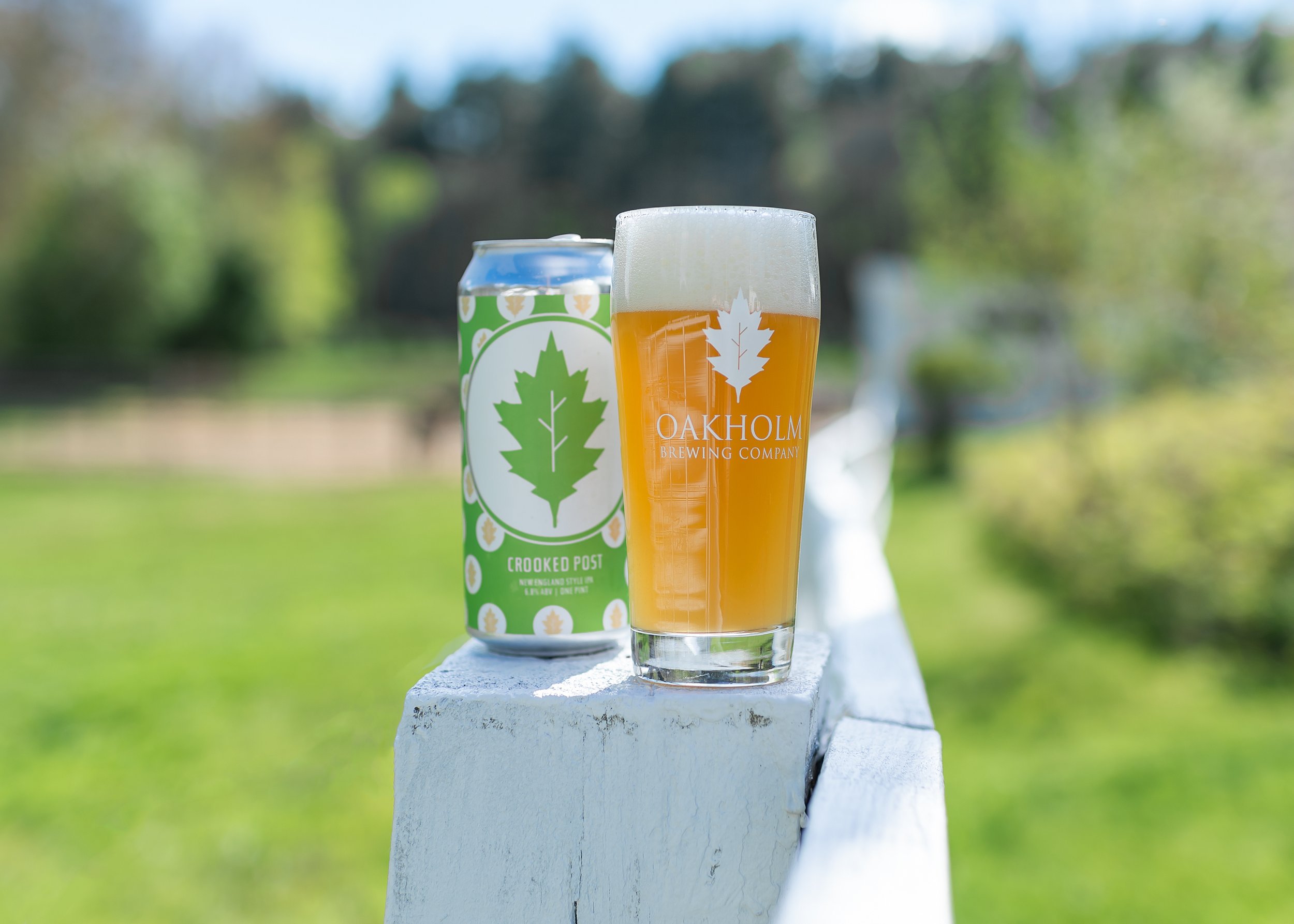 A can of Crooked Post beer and a glass of beer on a white painted wooden railing outdoors with greenery in the background.