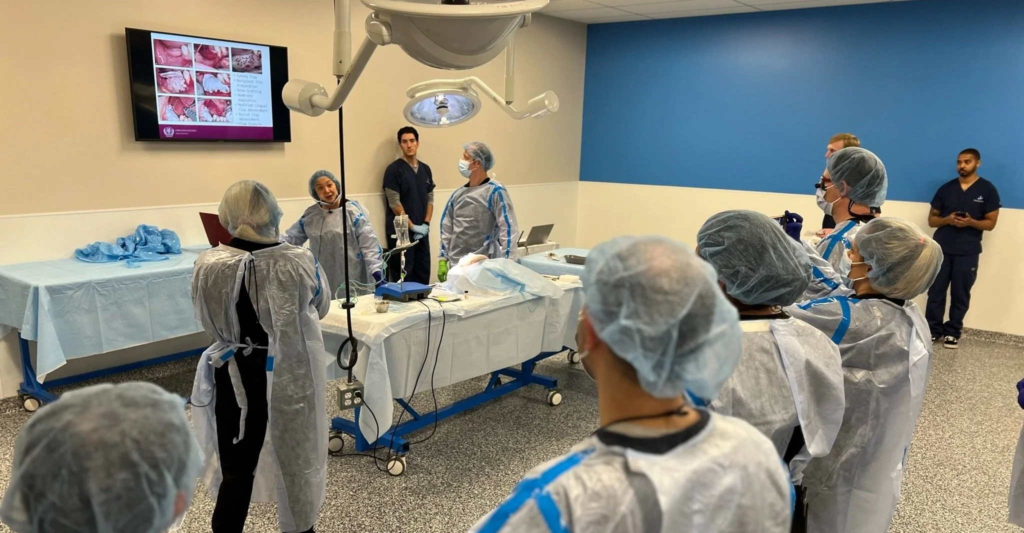 A team of dentists in surgical scrubs and protective gear attending a training or demonstration in a hospital room with a large monitor displaying dental images.