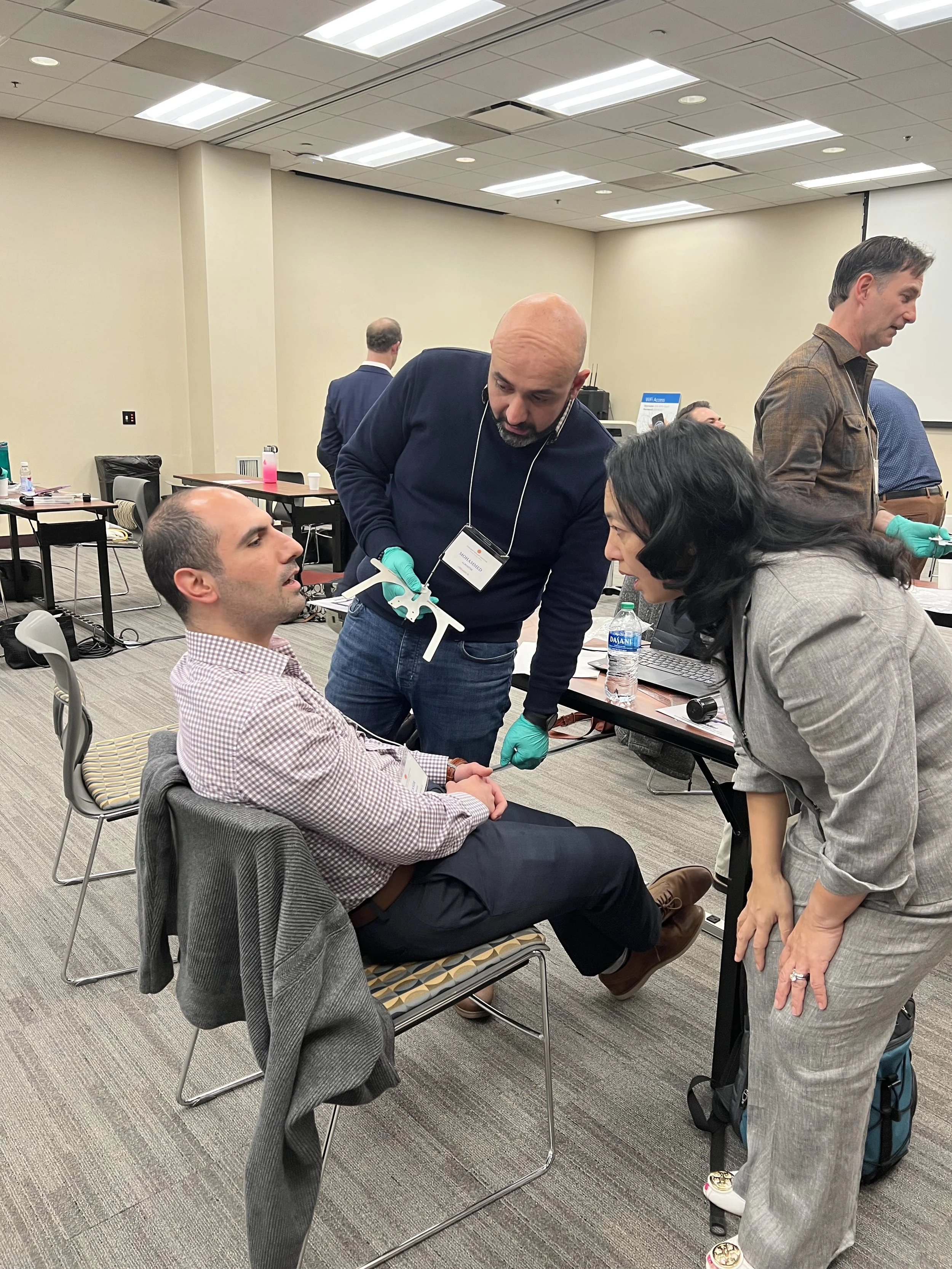 A dental professional conducting an oral exam on a seated man, with a woman leaning in to observe at a training session.