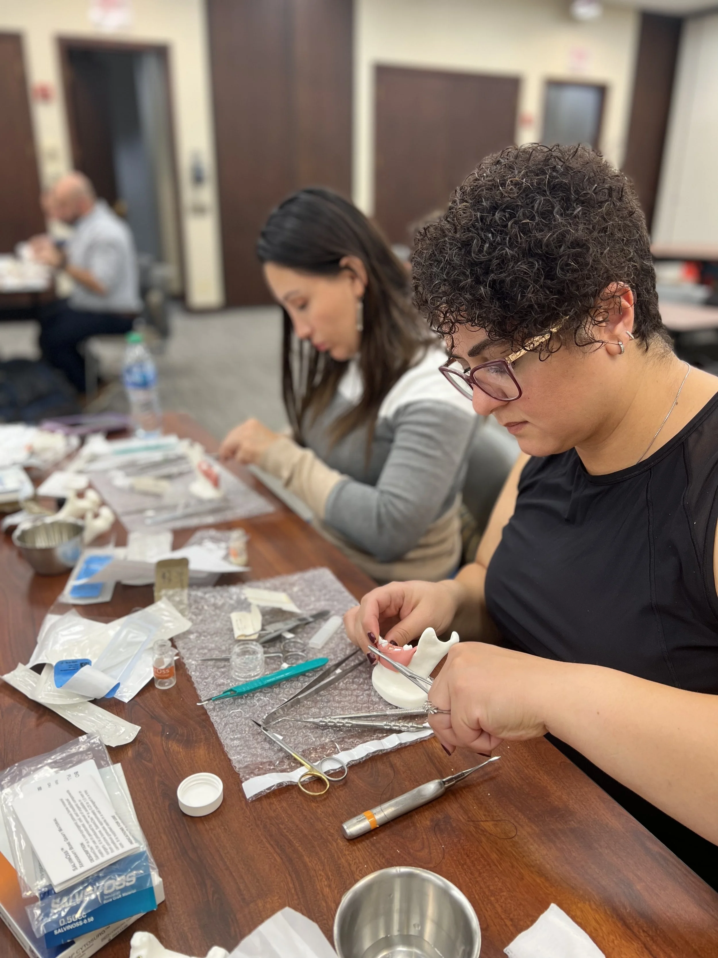 Two women working on dental prosthetics at a table with dental tools, models, and supplies, in a classroom setting.