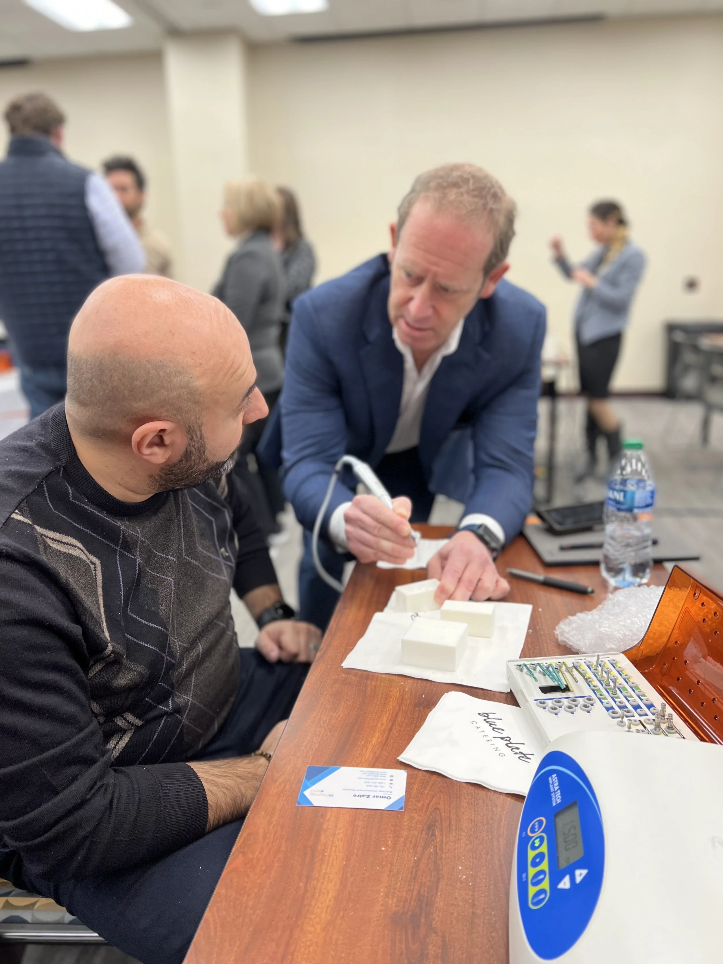 A man with a shaved head and beard is seated at a table, engaging in a conversation with an older man in a blue suit. The older man is holding device and demonstratingThe table has various items, including dental tools, a water bottle, and a digital scale.