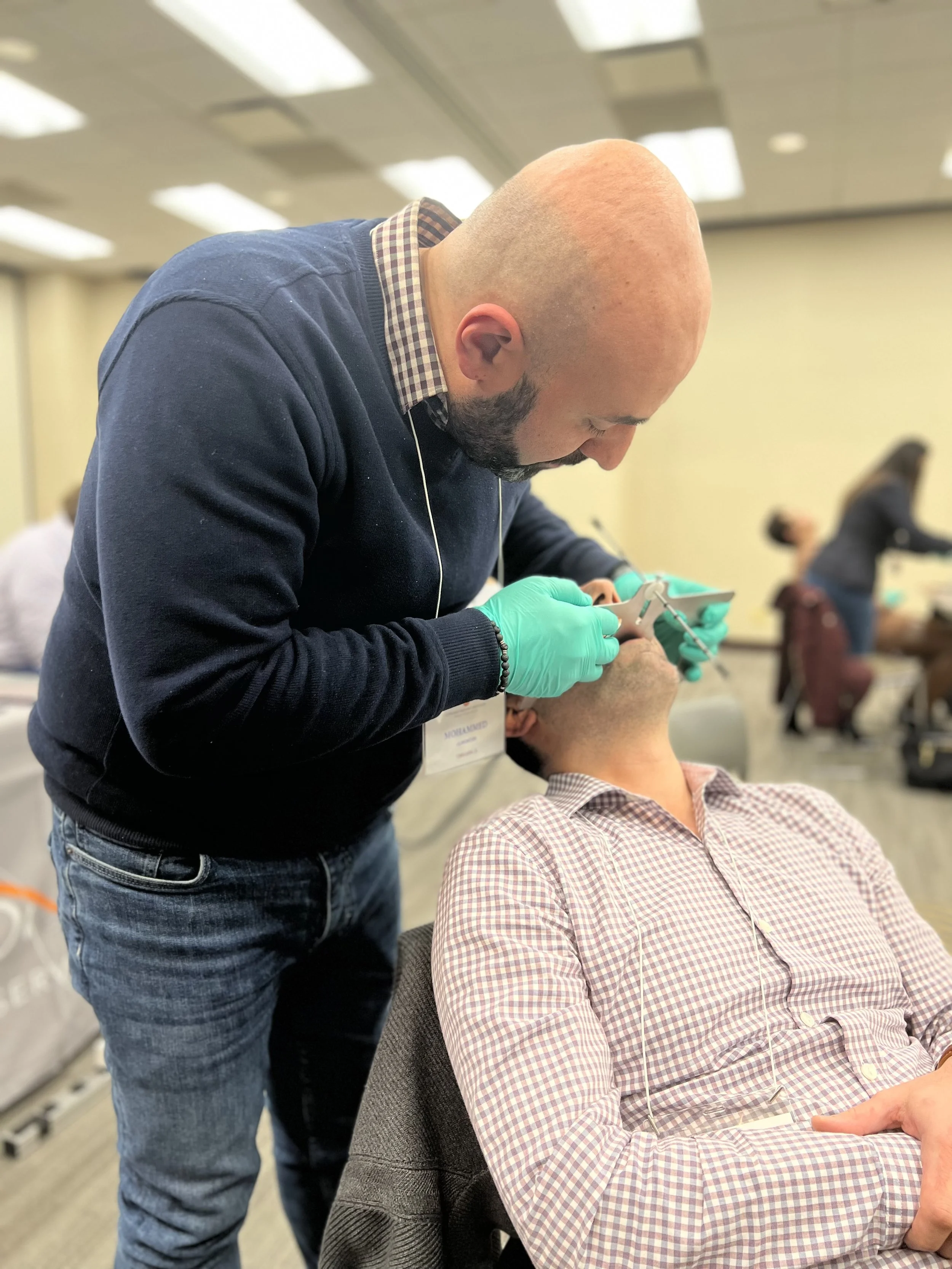 A man with a bald head and beard, wearing a dark sweater and green gloves, is administering a dental injection to another man sitting in a chair, who is wearing a checkered shirt in a busy indoor setting.