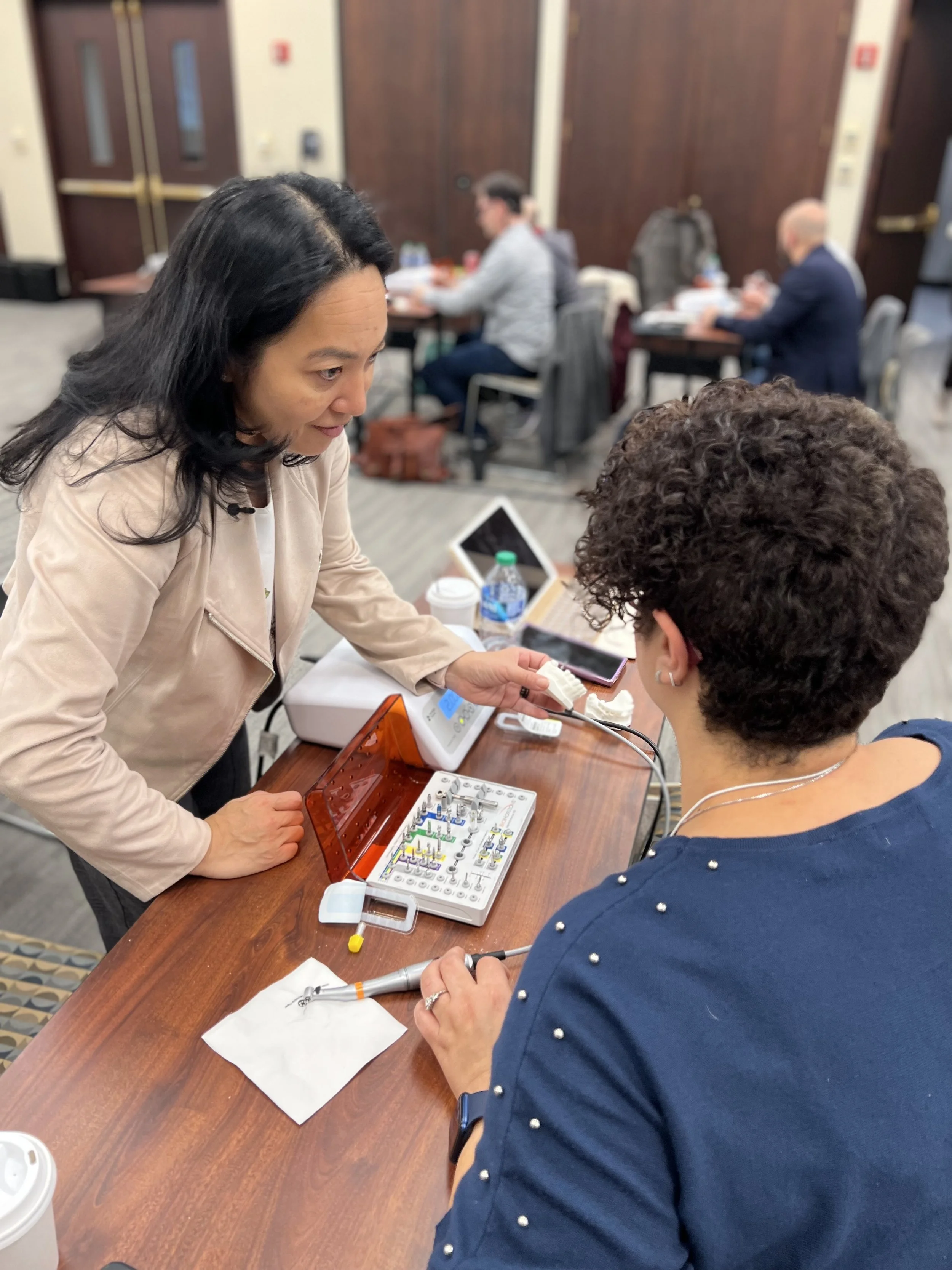 Two women at a table, one demonstrating a dental device to the other, with a group of people studying in the background.