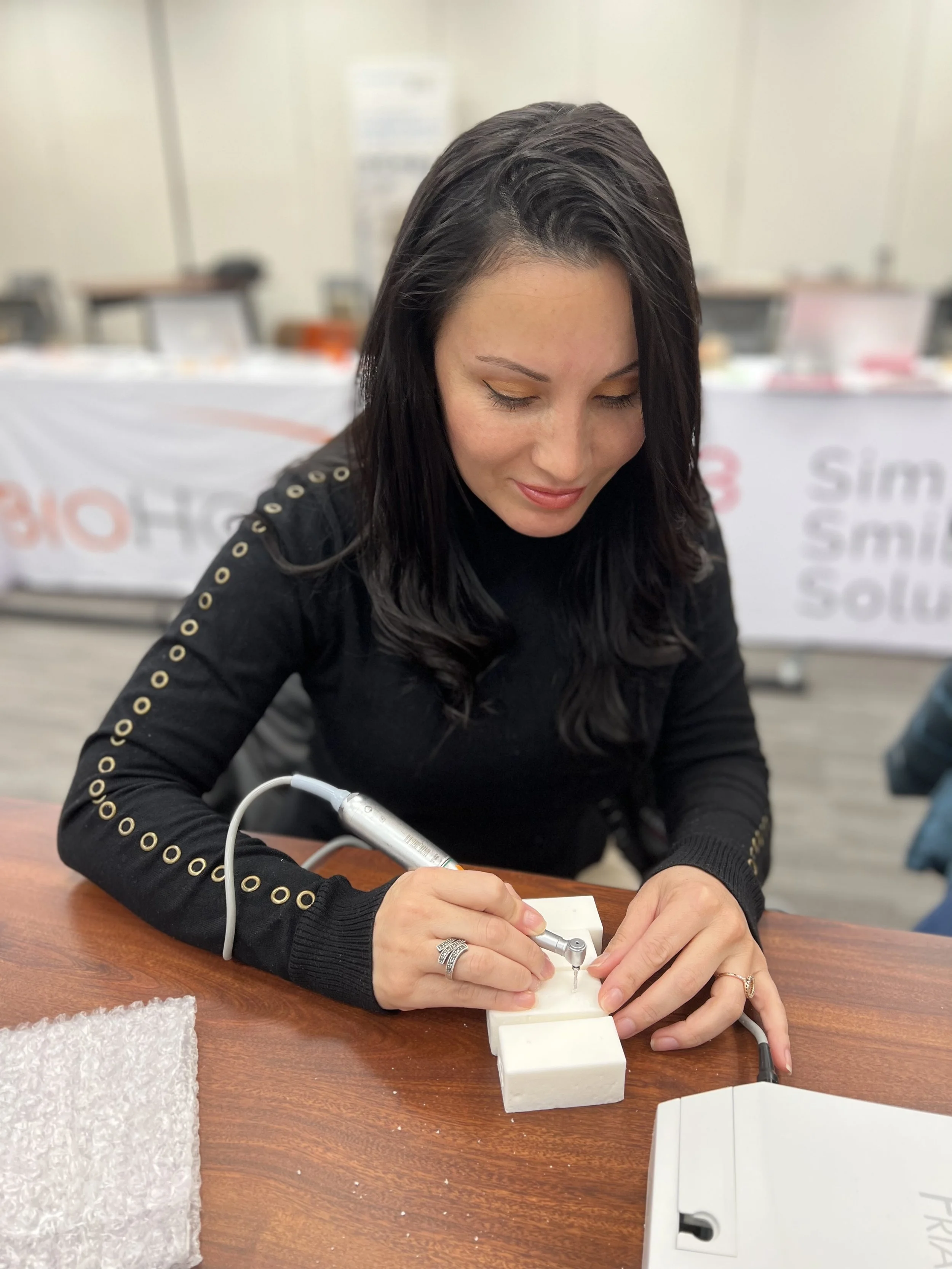 A woman with black hair and a black sweater with gold eyelets along the sleeves is working with a dental milling machine on a small white block at a table in a classroom setting.