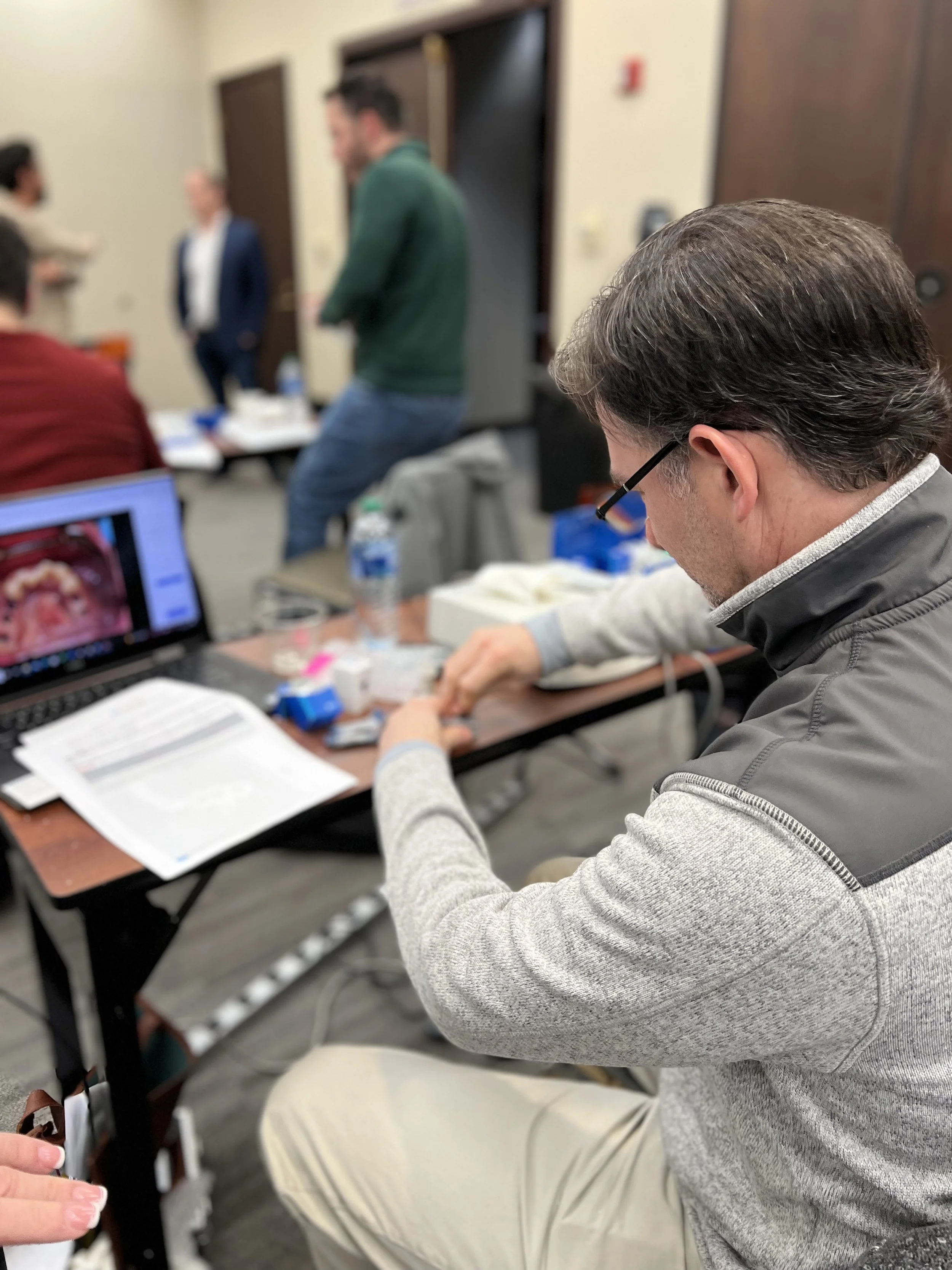 A dentist with glasses and a gray jacket seated at a table, looking at dental images on a laptop, with other people standing and talking in the background.