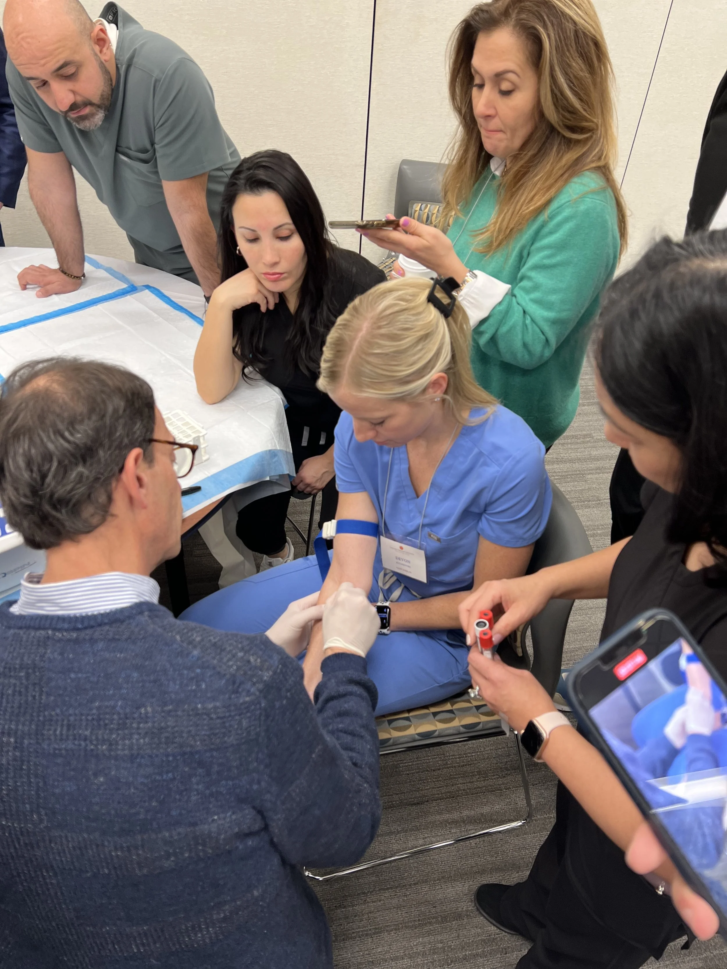 A dentist draws blood from a woman's arm while others observe and record the procedure at The Implant Method training.
