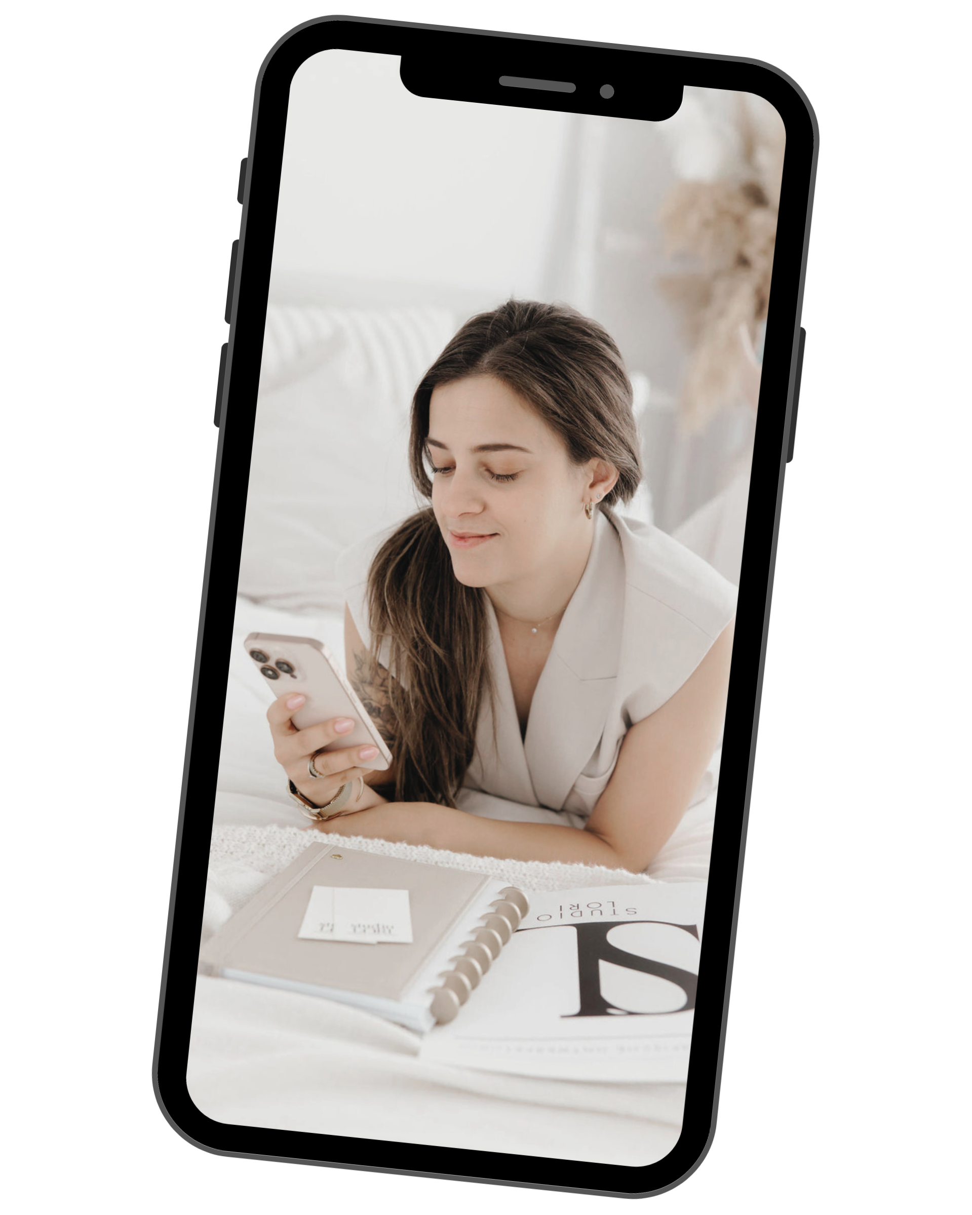 A woman lying on a bed, looking at her phone with a white and beige desk planner in front of her, in a bright, cozy room.