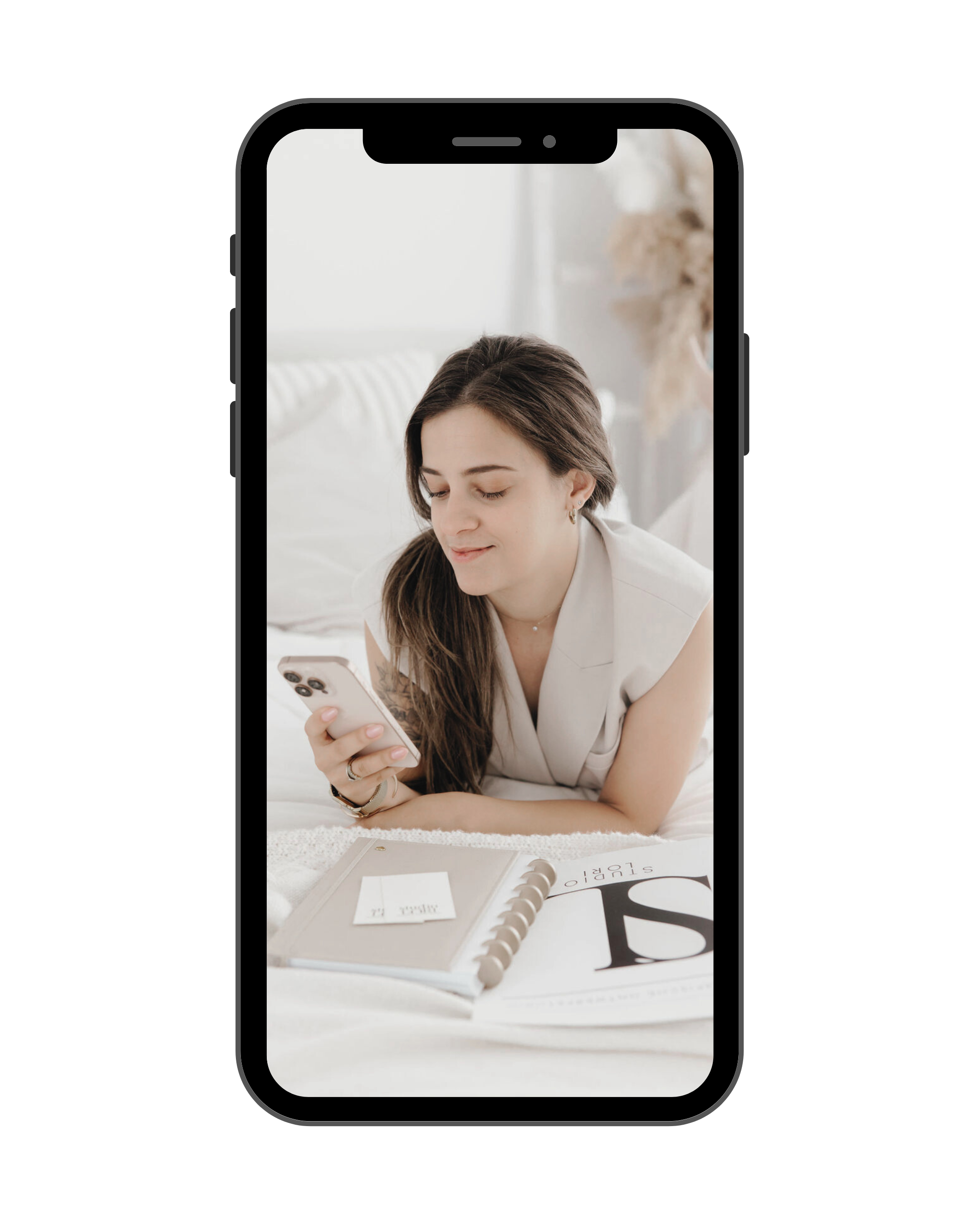 A woman lying on her stomach on a bed, looking at her phone with a slight smile, surrounded by a notebook, magazine, and papers.