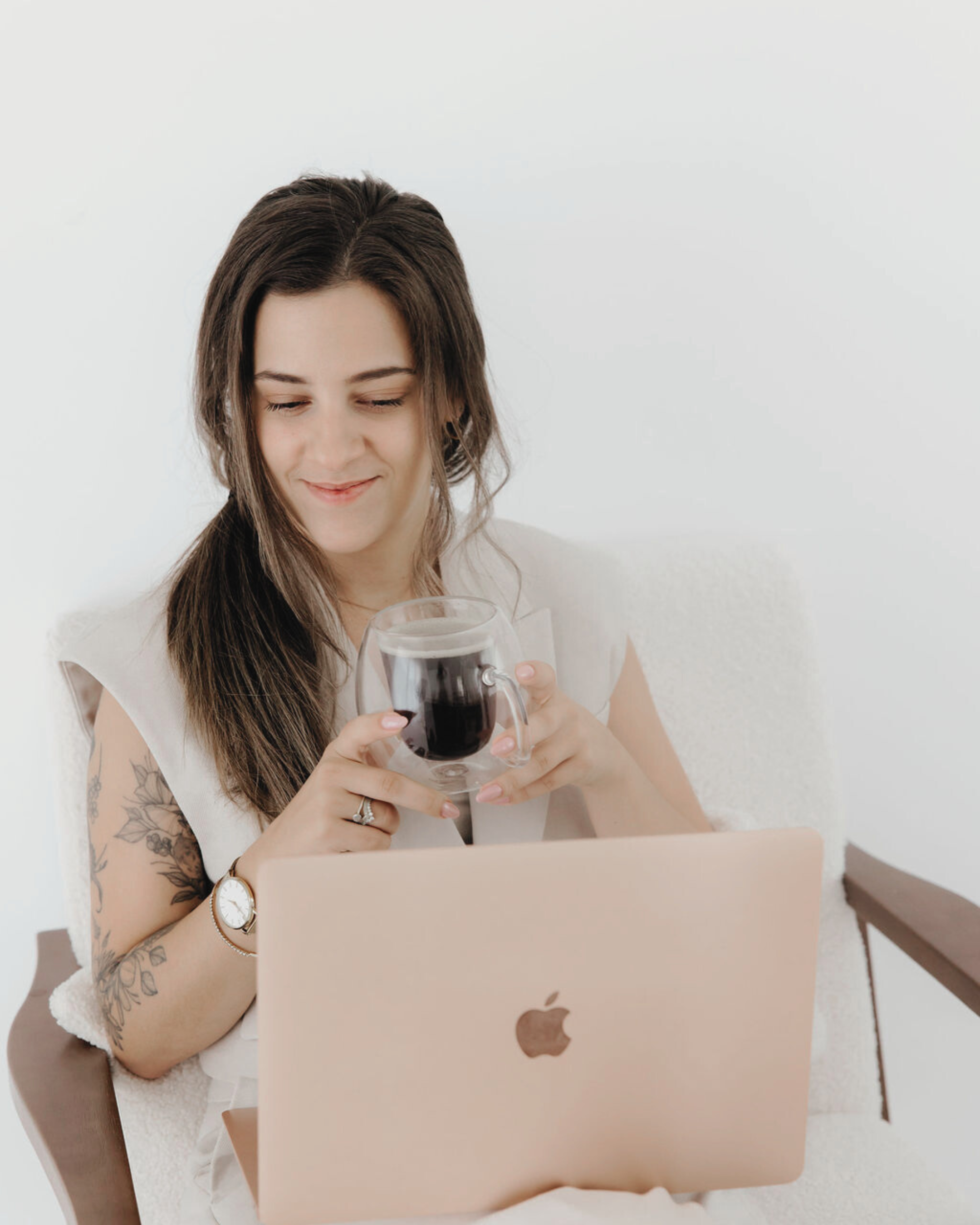 A woman with long brown hair, tattoos, and a wristwatch, smiling and sitting on a white chair with a beige laptop on her lap, holding a glass mug of dark coffee, looking at her laptop.