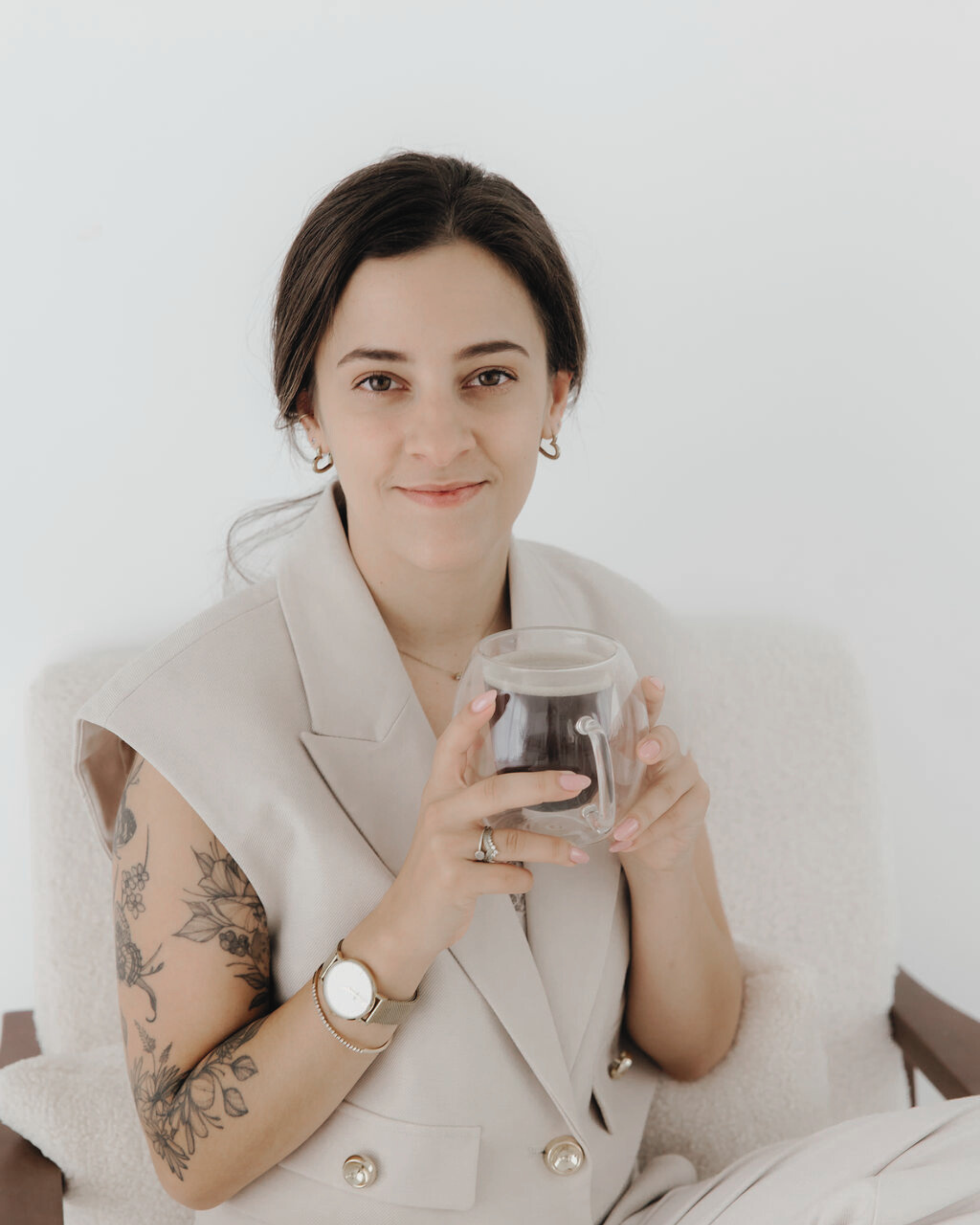 Woman with dark hair and tattoos on her arm, holding a clear mug with dark liquid, sitting on a light-colored armchair against a plain background.