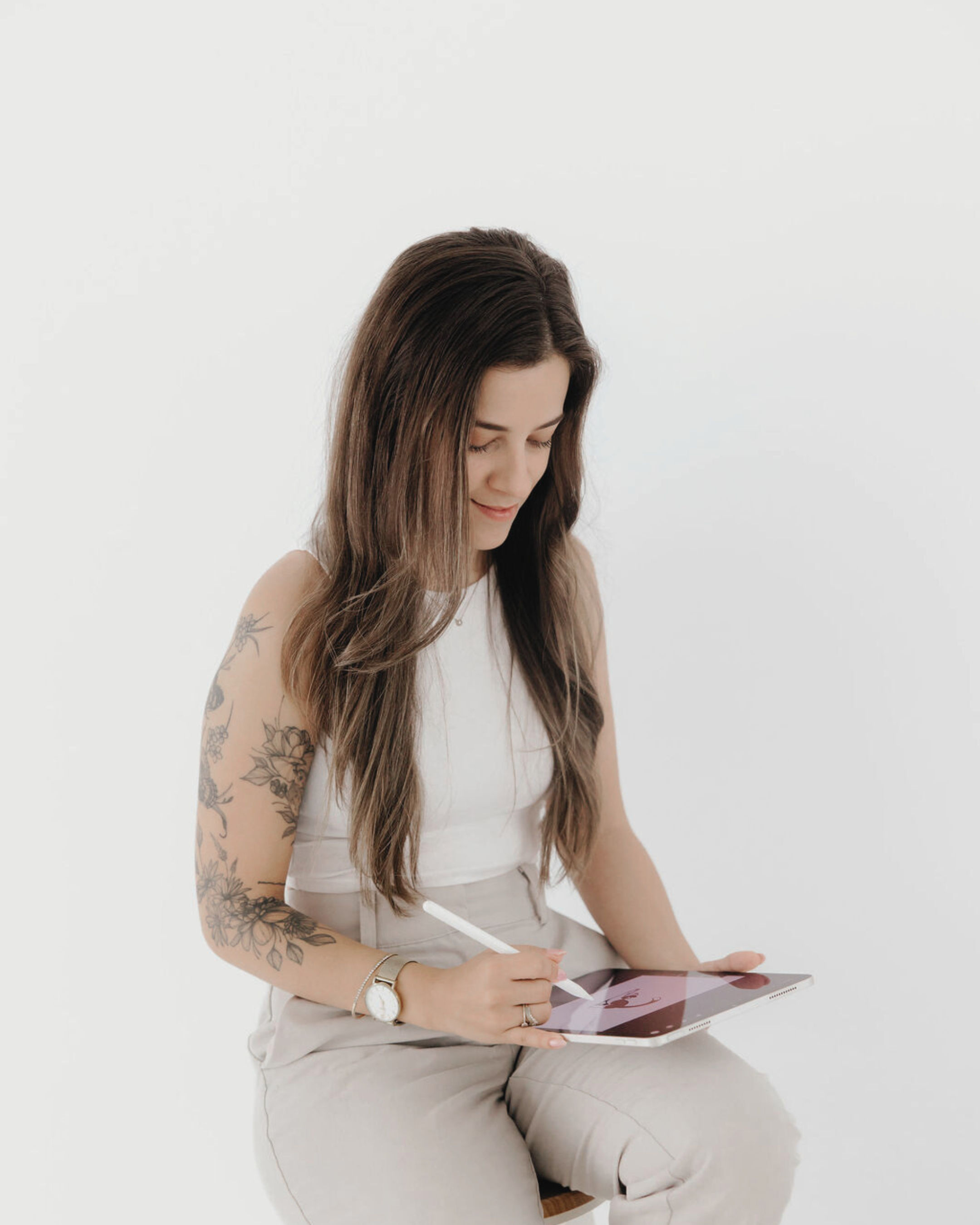 A woman with long brown hair and tattoos on her left arm, sitting and using a stylus on a tablet with a pink drawing on the screen.