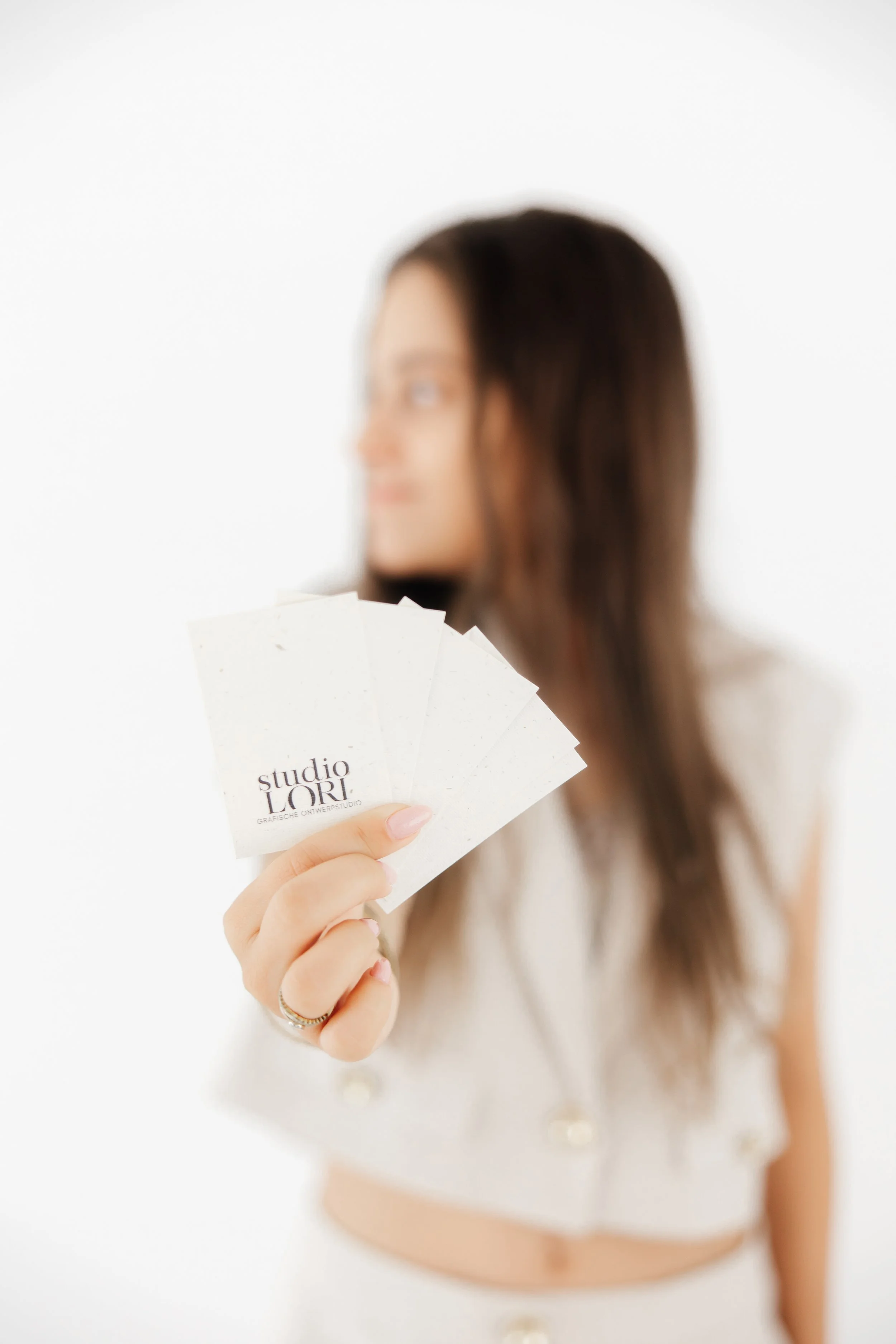 A woman holding five business cards with the text "studio LORI" in focus, while her face and body are out of focus in the background. Designing geboortekaartjes, geboortekaartjes op maat, branding, logo