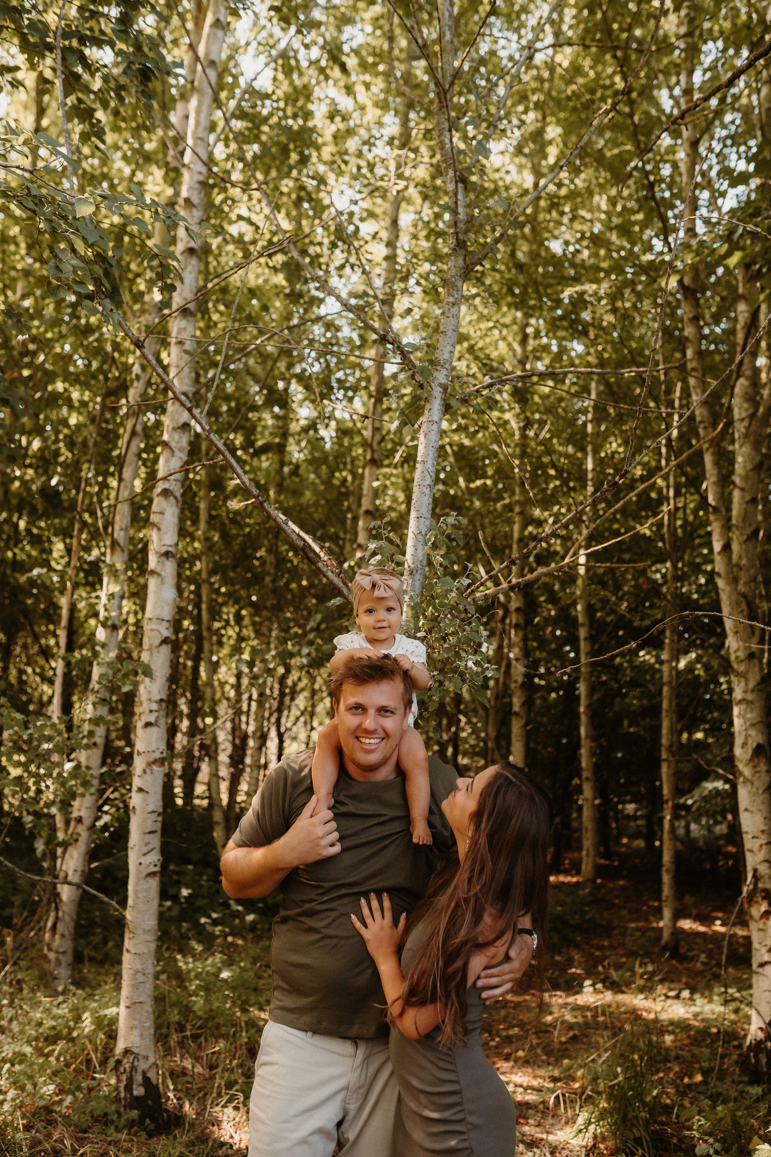 A family of three in a forest, with the father carrying the young daughter on his shoulders, and the mother looking up at her. The sunlight filters through the trees.