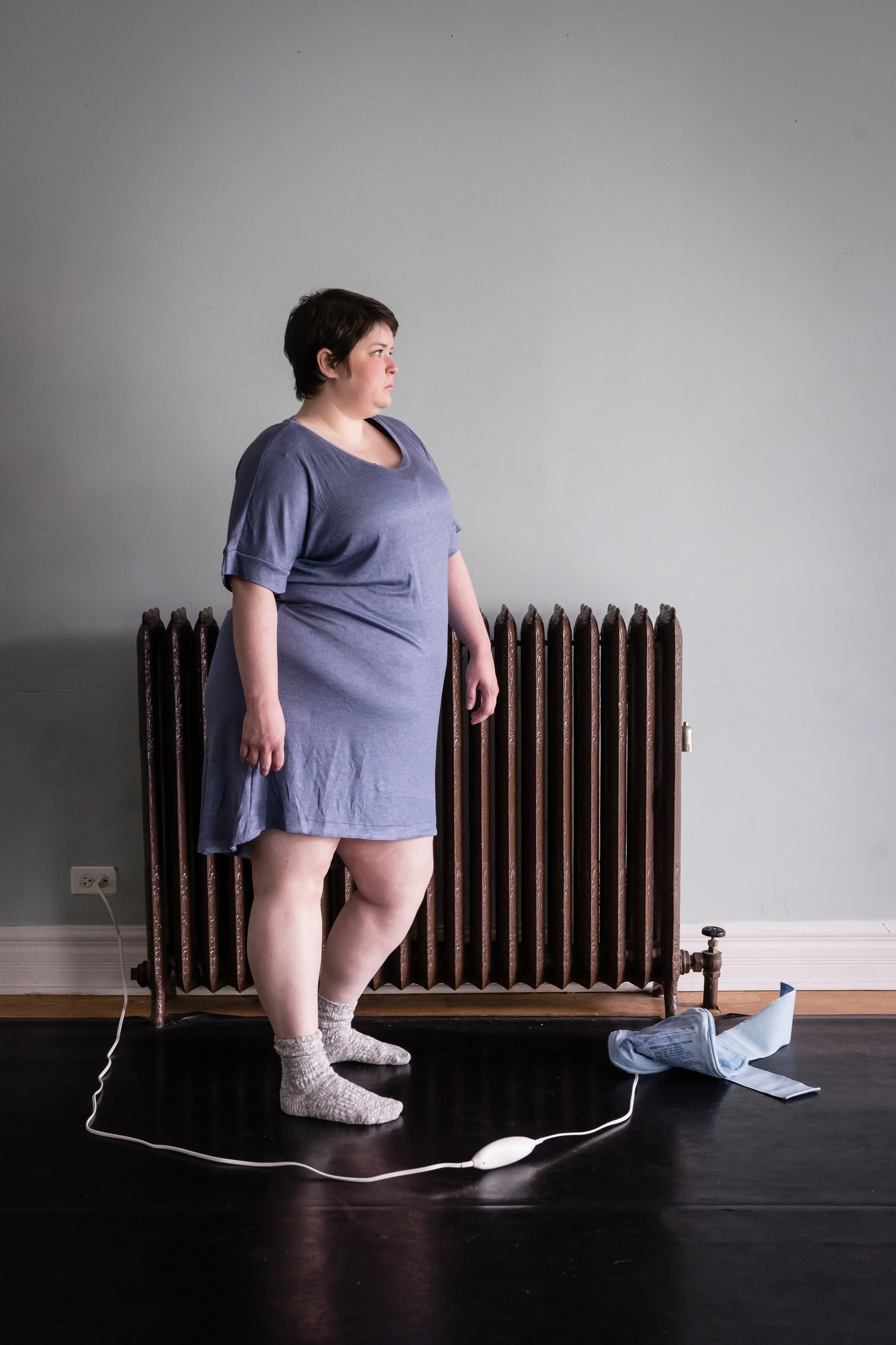 Maggie stands in front of an ornate brown radiator wearing a light blue nightgown and grey socks. She faces right, bending her back knee and letting that hip drop. A blue heating pad lays on the floor in front of her, the cord circling around her.