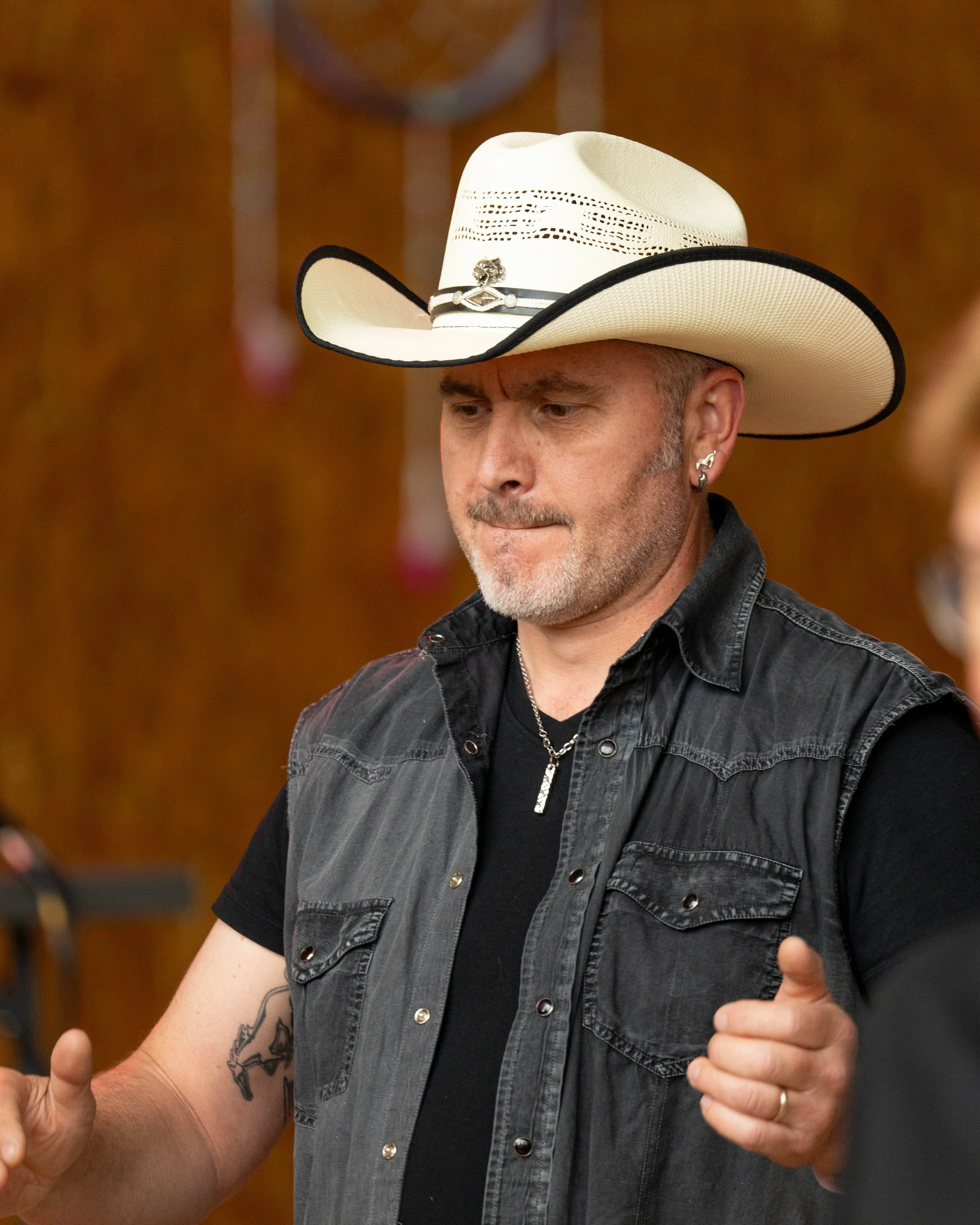 Homme portant un chapeau de cowboy blanc, une veste en denim gris foncé, une chaîne en collier, et une bague, dans un décor intérieur.