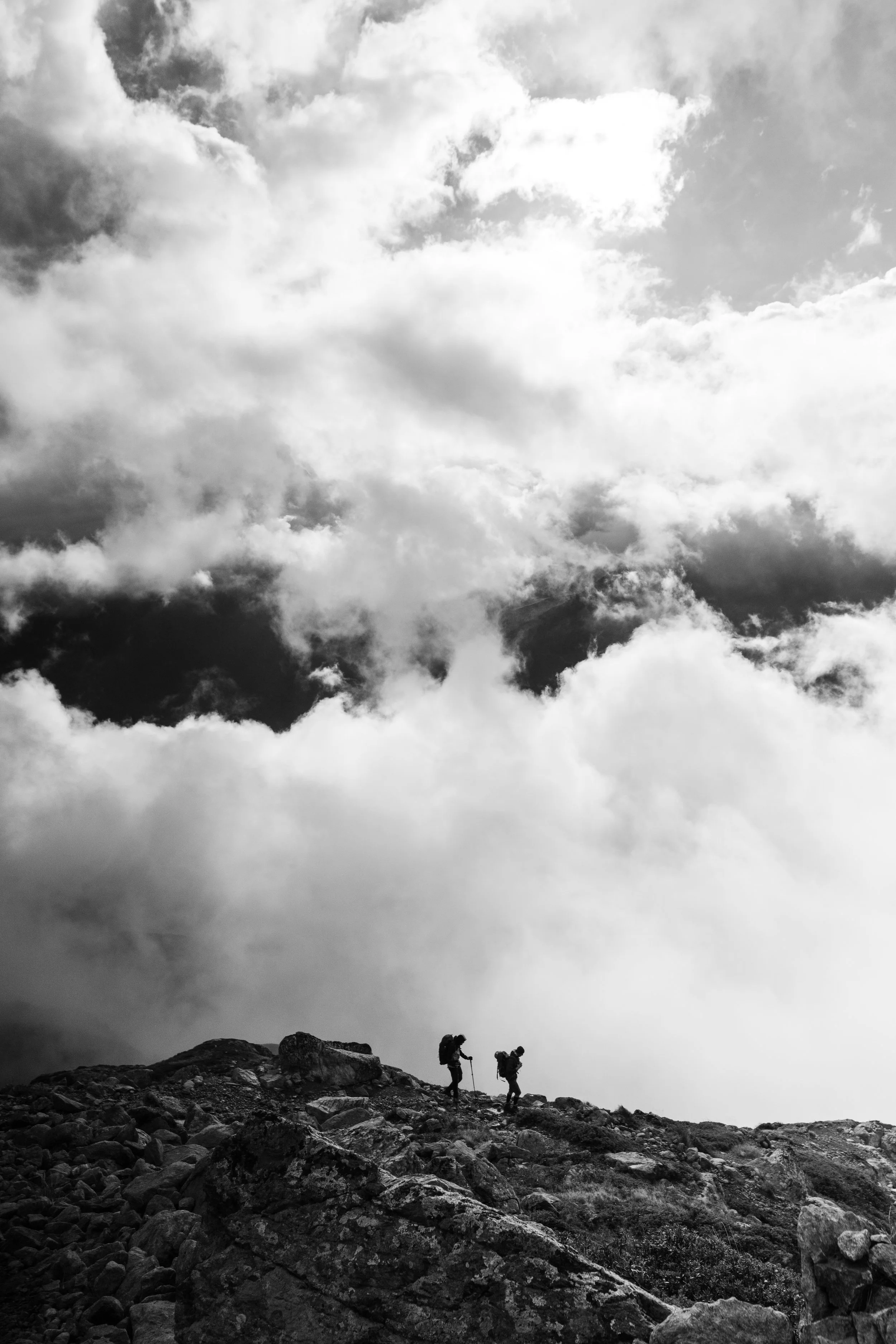Deux randonneurs (Antonin et Romuald) durant le projet "Subir pour Soutenir" portant des sacs à dos et des bâtons de marche sur un terrain rocheux en montagne, sur le GR TMB, avec un ciel nuageux au-dessus.