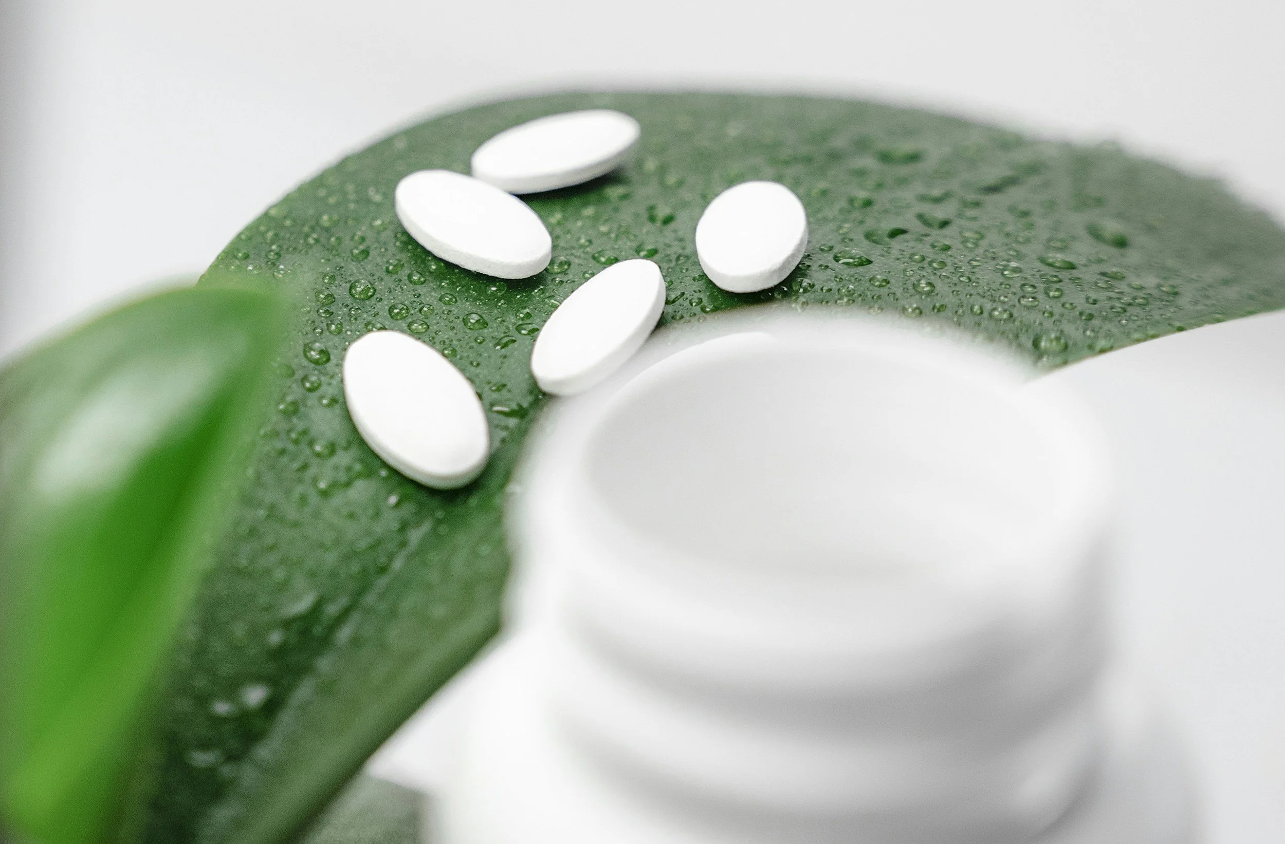 White oval pills on a green leaf with water droplets and a white container in the background.