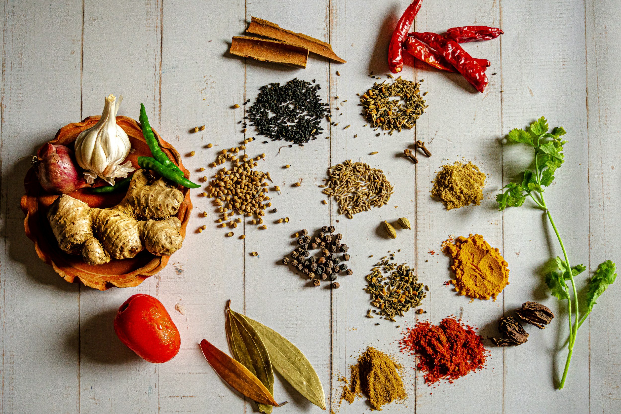 Various spices and herbs arranged on a white wooden surface, including garlic, ginger, green chili, dried red chili, turmeric, coriander, cumin, black pepper, and bay leaves, with a basket containing additional garlic, ginger, and onion.
