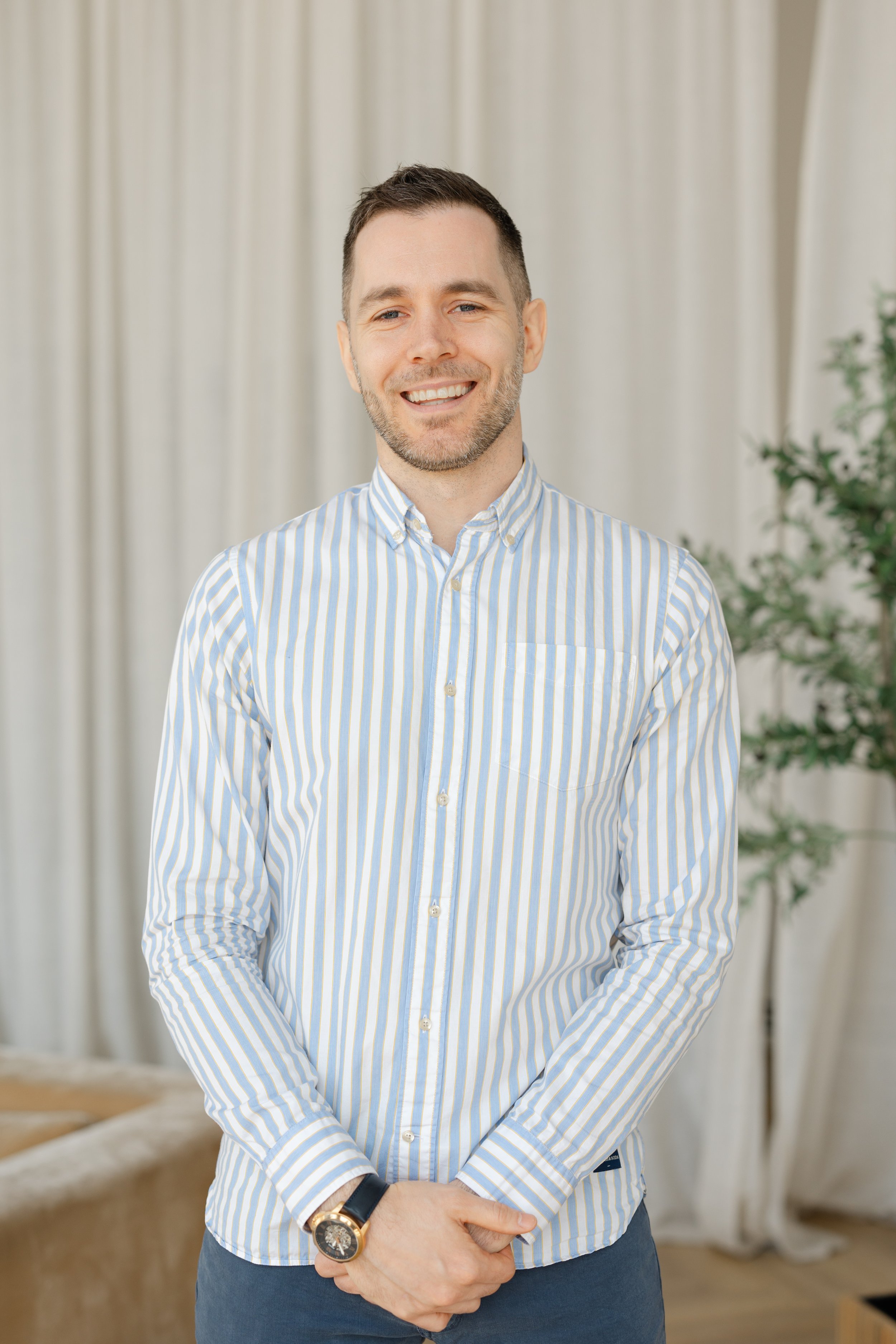 A smiling man in a light blue and white striped button-up shirt standing indoors with curtains and a plant behind him.