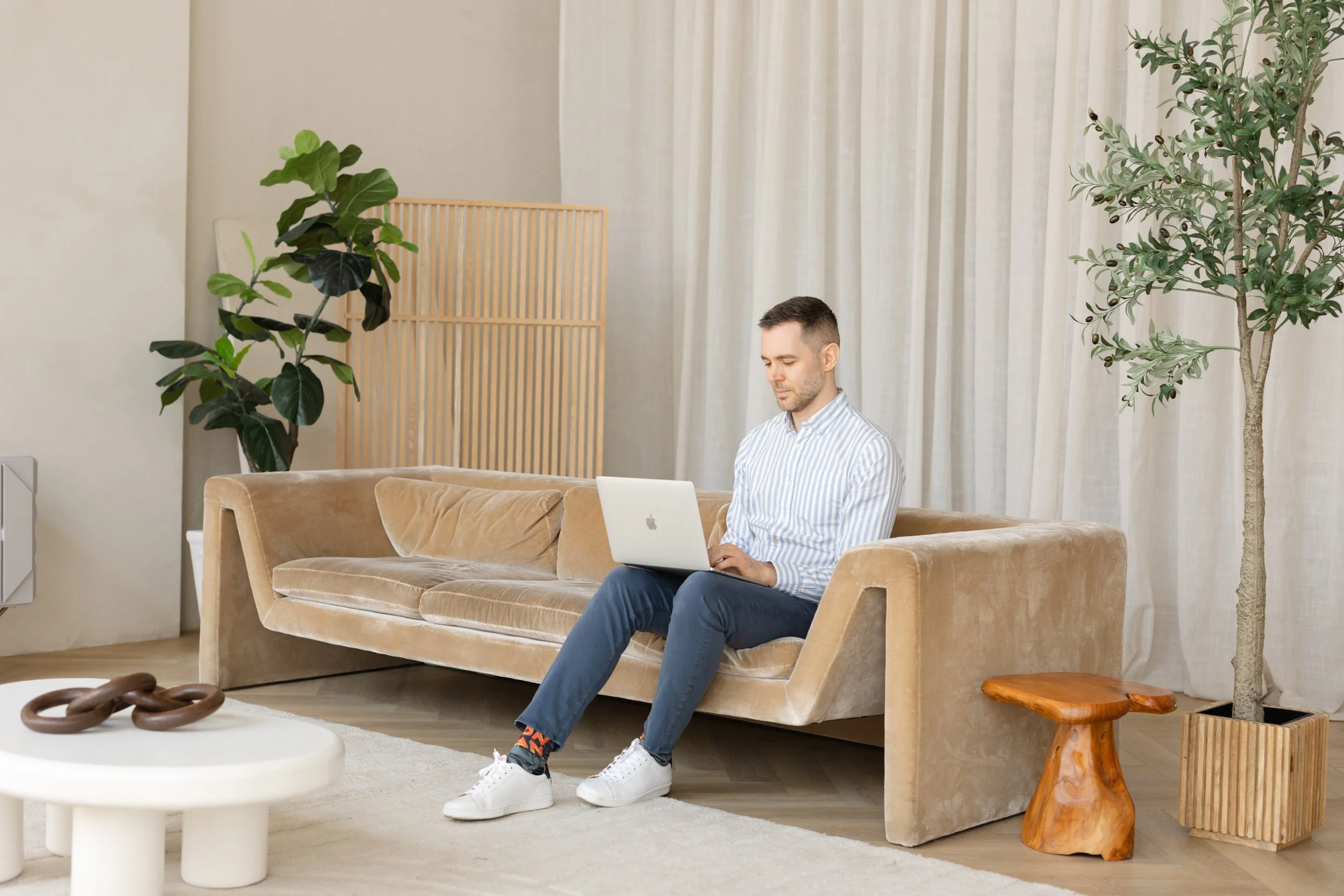 A man sitting on a beige velvet sofa working on a silver laptop in a living room with light-colored walls, large green plants, a wooden side table, and a white coffee table with an abstract sculpture.