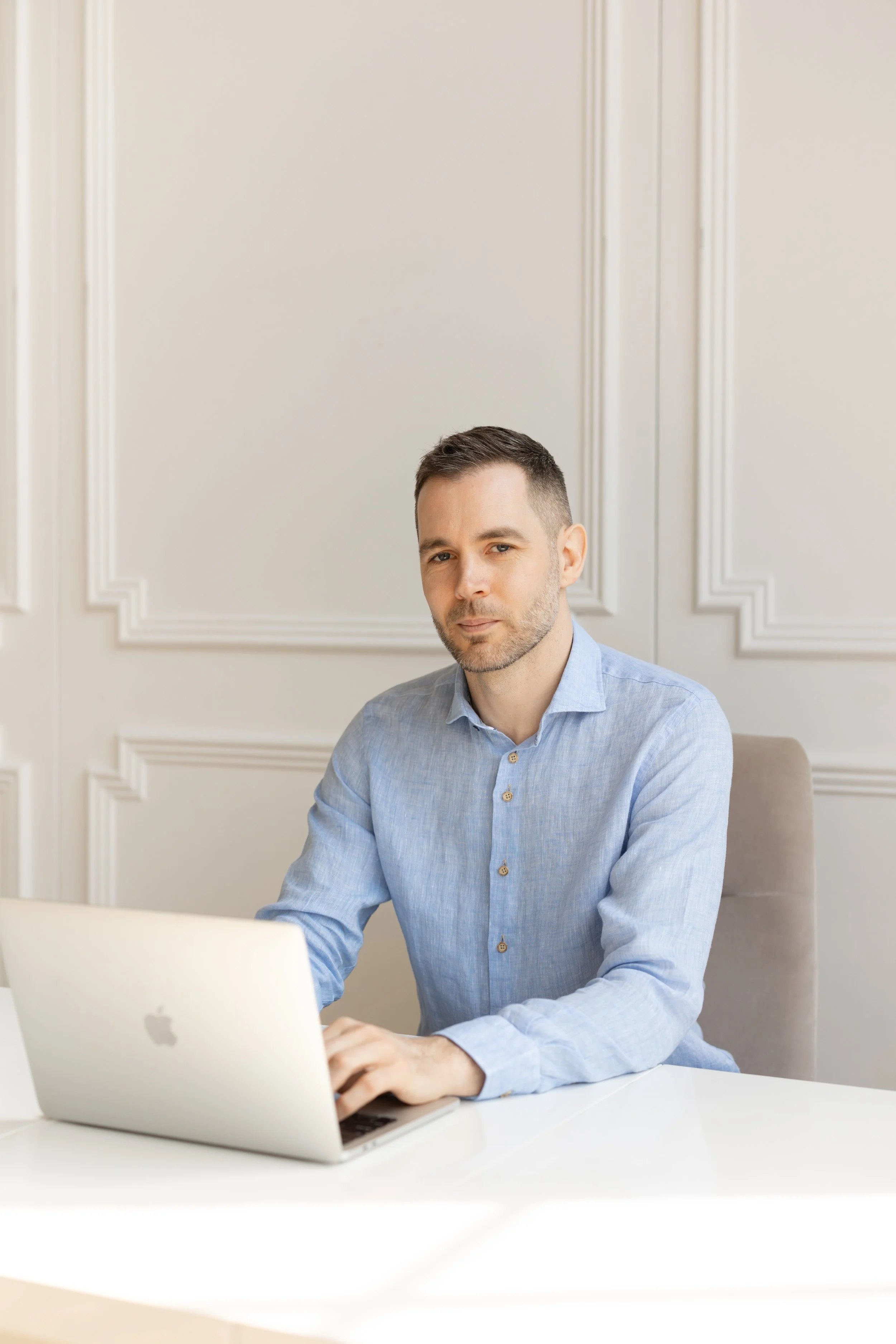 A man with short dark hair and a light beard, wearing a light blue button-up shirt, sits at a white desk with an open silver laptop, in a room with cream-colored walls with decorative molding.