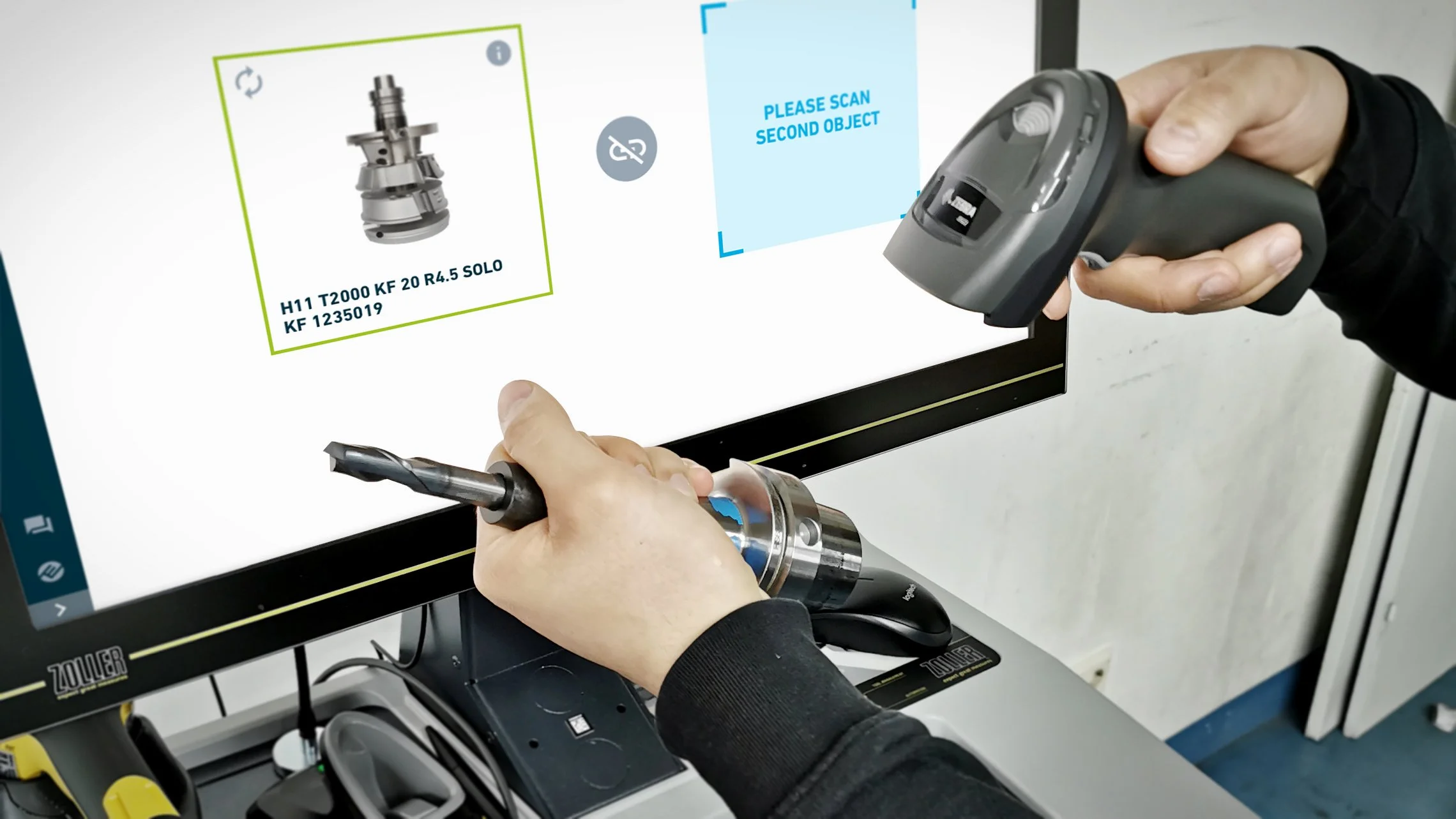 Person scannt ein Werkzeug und einen Teil mit einem Handscanner vor einem Computerbildschirm in einem technischen Umfeld.