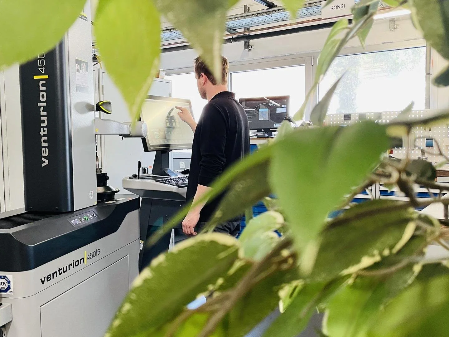 Mann arbeitet an einem wissenschaftlichen Gerät in einem Labor, im Hintergrund ein Computerbildschirm und Pflanzen im Vordergrund