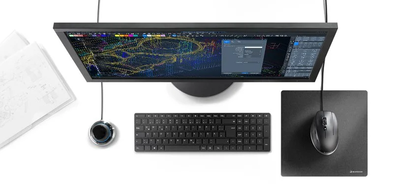 Ein Desktop-Computer mit einem großen Monitor, einer Tastatur, einer Maus auf einer schwarzen Unterlage, einer Webcam und einem Notizbuch mit technischen Zeichnungen.