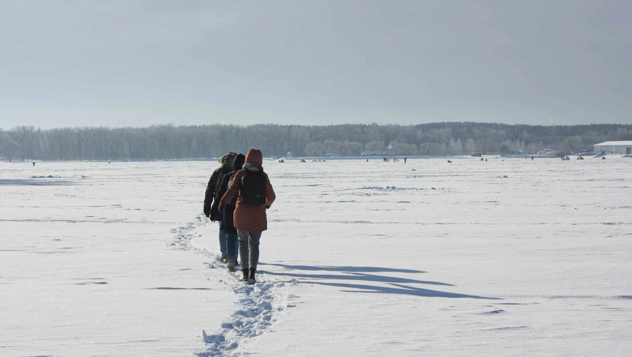 Four people walking in a line across a snow-covered frozen lake, with a distant tree line and structures in the background, on a clear winter day.