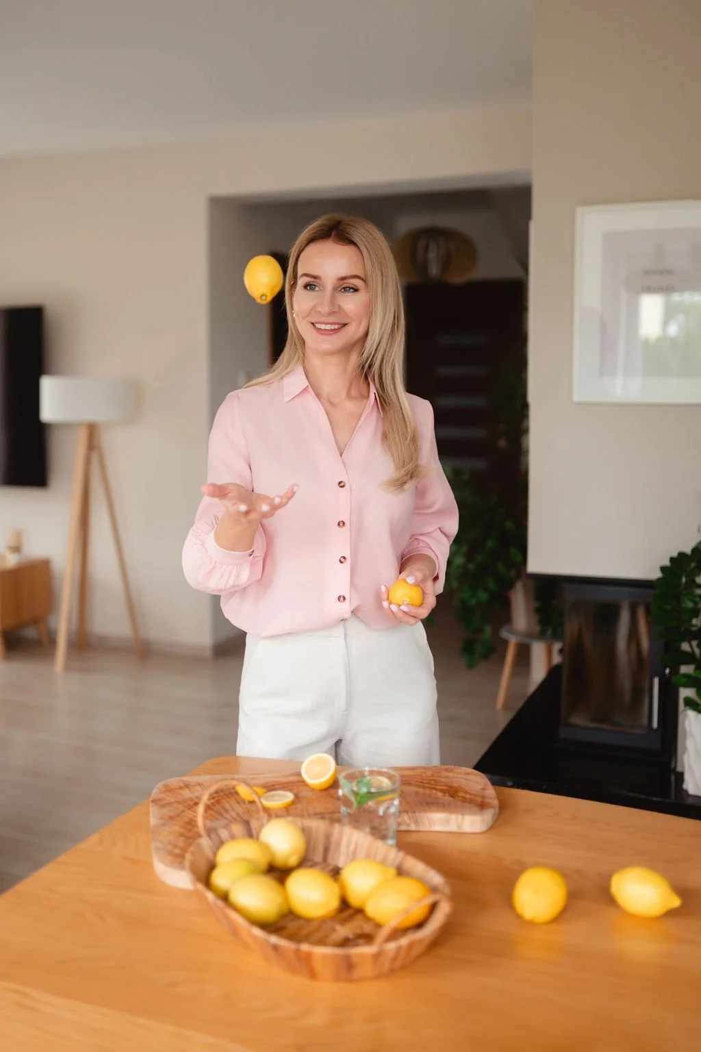 A woman in a pink blouse and white pants smiling and juggling a lemon in a modern home kitchen with lemons and a glass of water on the table.