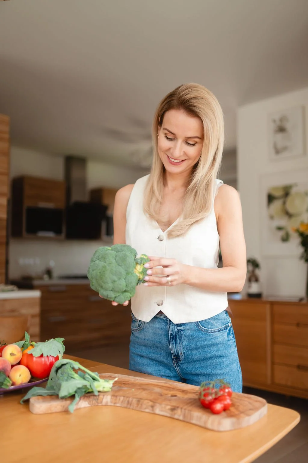 A woman in a sleeveless white top and blue jeans holds a head of broccoli while standing in a kitchen with wooden cabinets. There are tomatoes, apples, and broccoli on the wooden table in front of her.